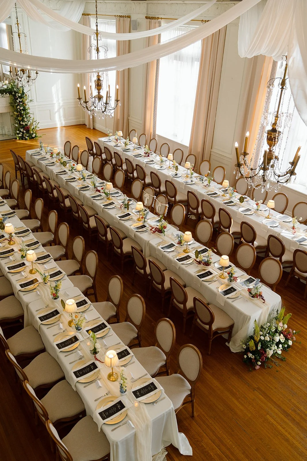 Elegant banquet hall decorated for a wedding with long white tables, floral centerpieces, small lamps, chandeliers, draped white curtains, and cream-colored chairs.