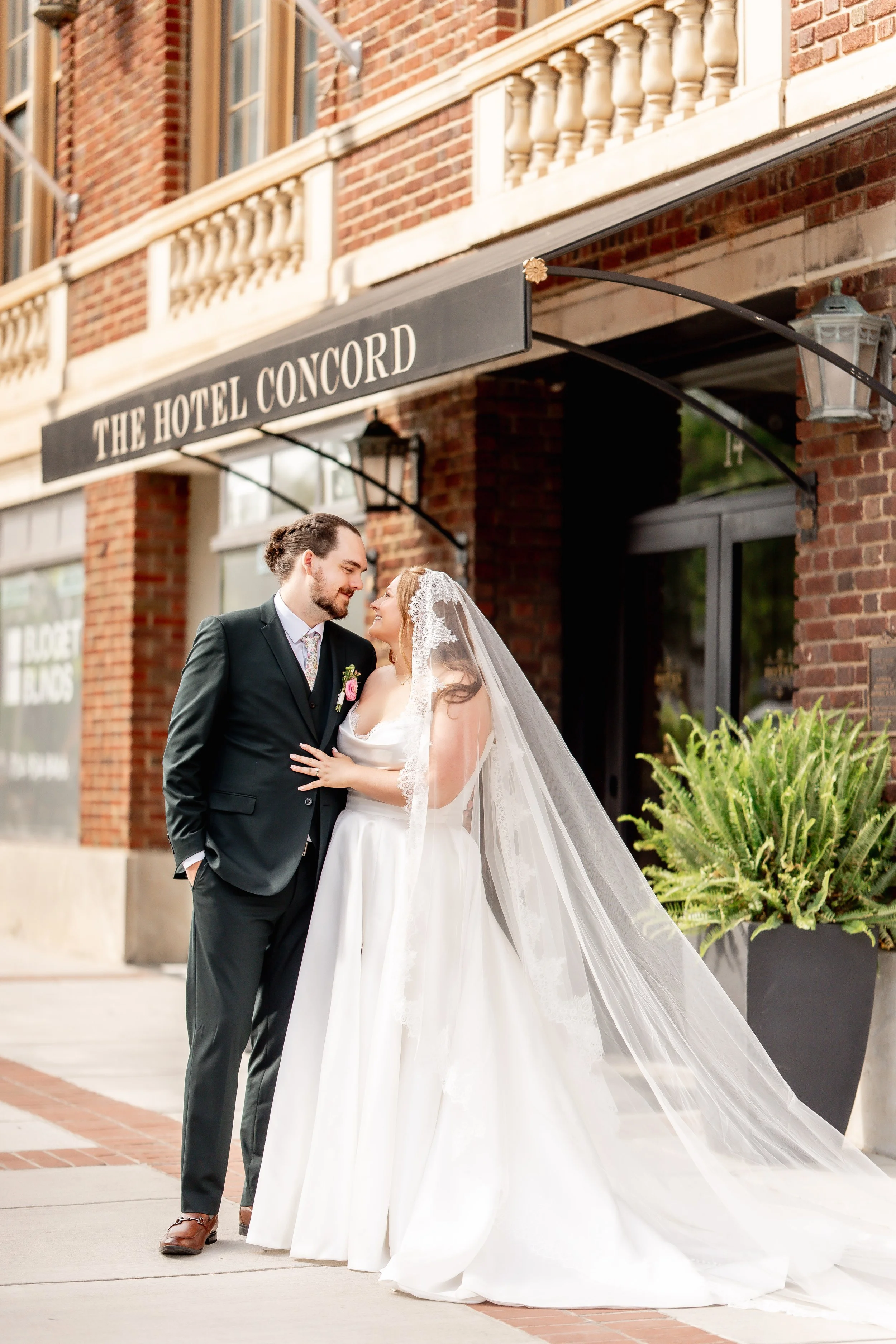 A bride and groom standing close together outside a brick building labeled 'The Hotel Concord'. The groom is in a black suit with a floral tie, and the bride is in a white wedding dress with a long veil. They are smiling at each other.