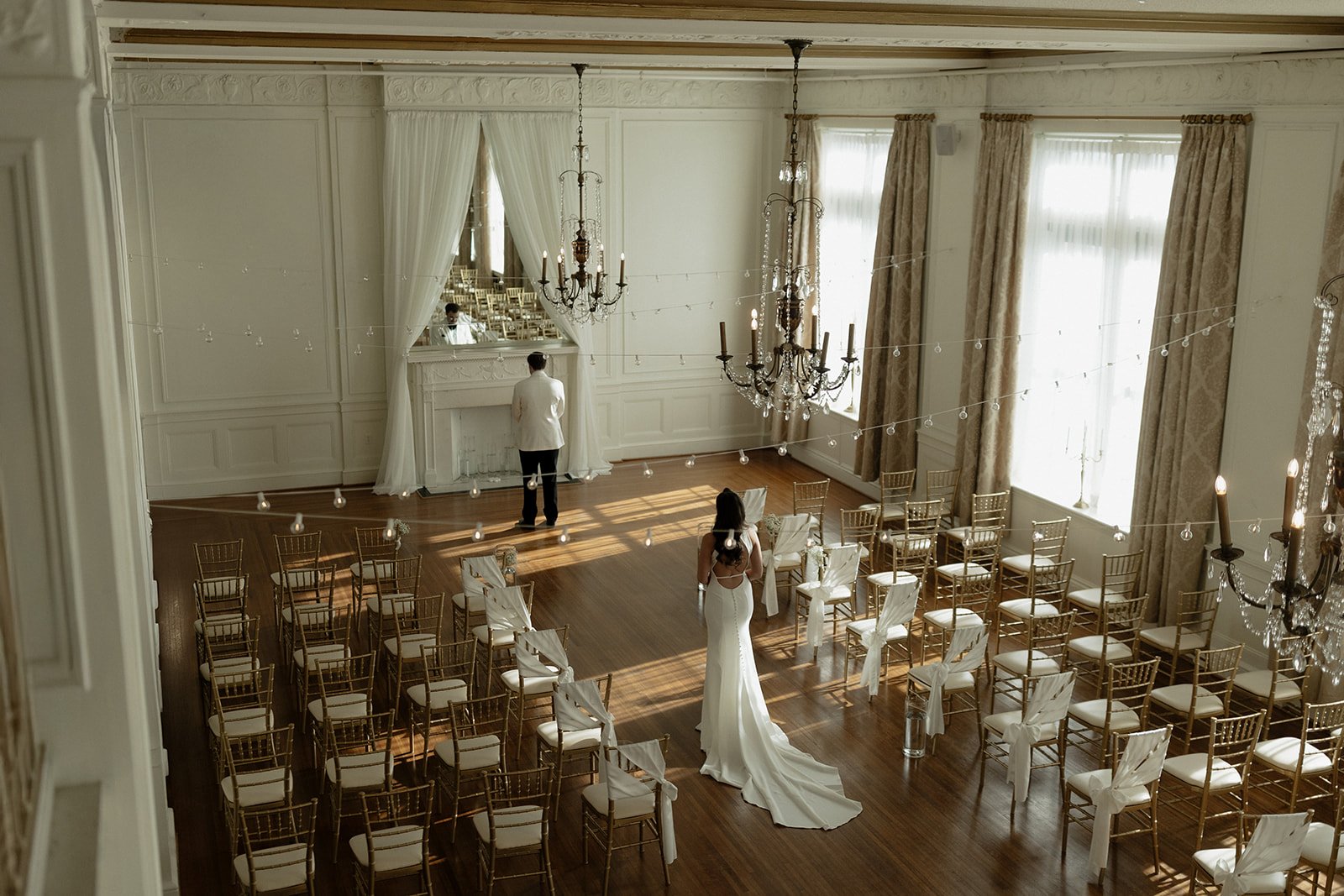 Elegant wedding ceremony setup in a bright, spacious room with gold chairs, a white fireplace with curtains, chandeliers, and large windows with curtains. A bride in a white gown stands near the chairs, and a groom in a white tuxedo looks at the fire