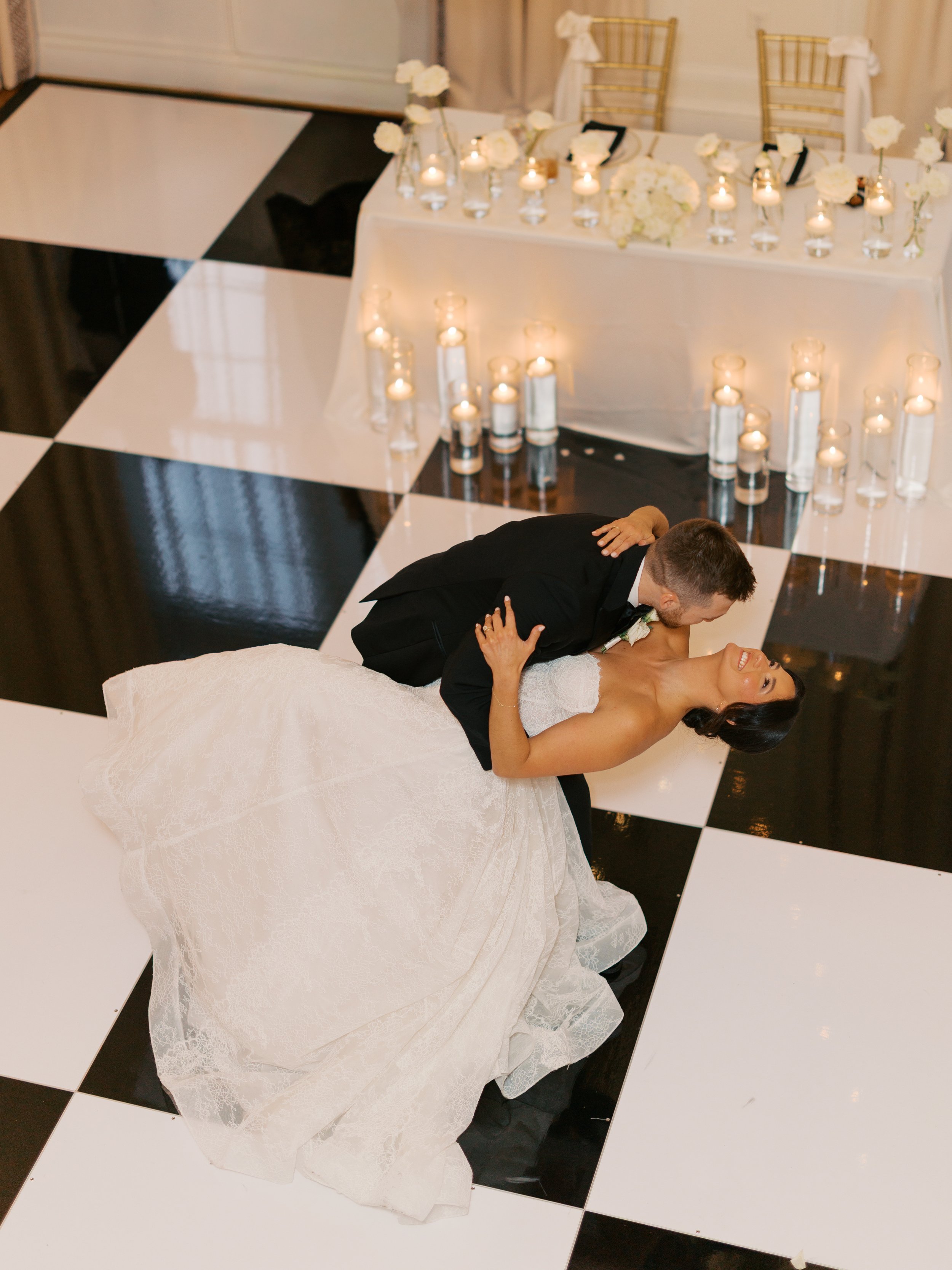 A newly married couple is sharing a kiss during their wedding reception, with a table decorated with candles, white flowers, and gift boxes behind them on a checkered black and white floor.