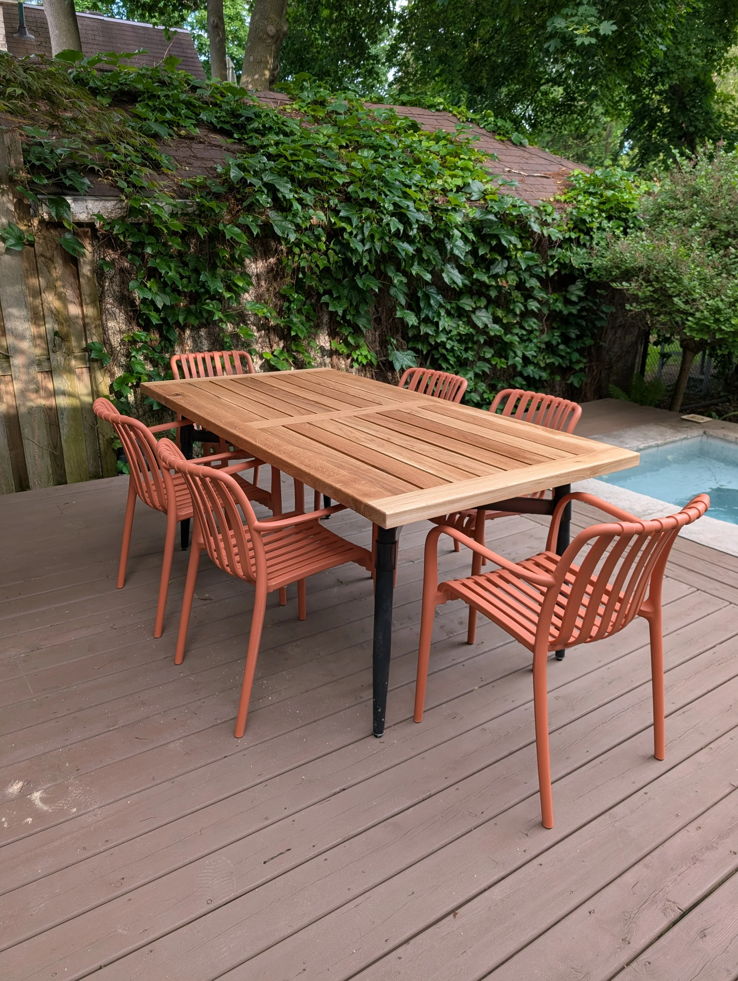 An outdoor patio with a wooden dining table surrounded by eight peach-colored chairs. A small pool is visible to the right, with lush green trees and ivy covering a shed wall in the background.