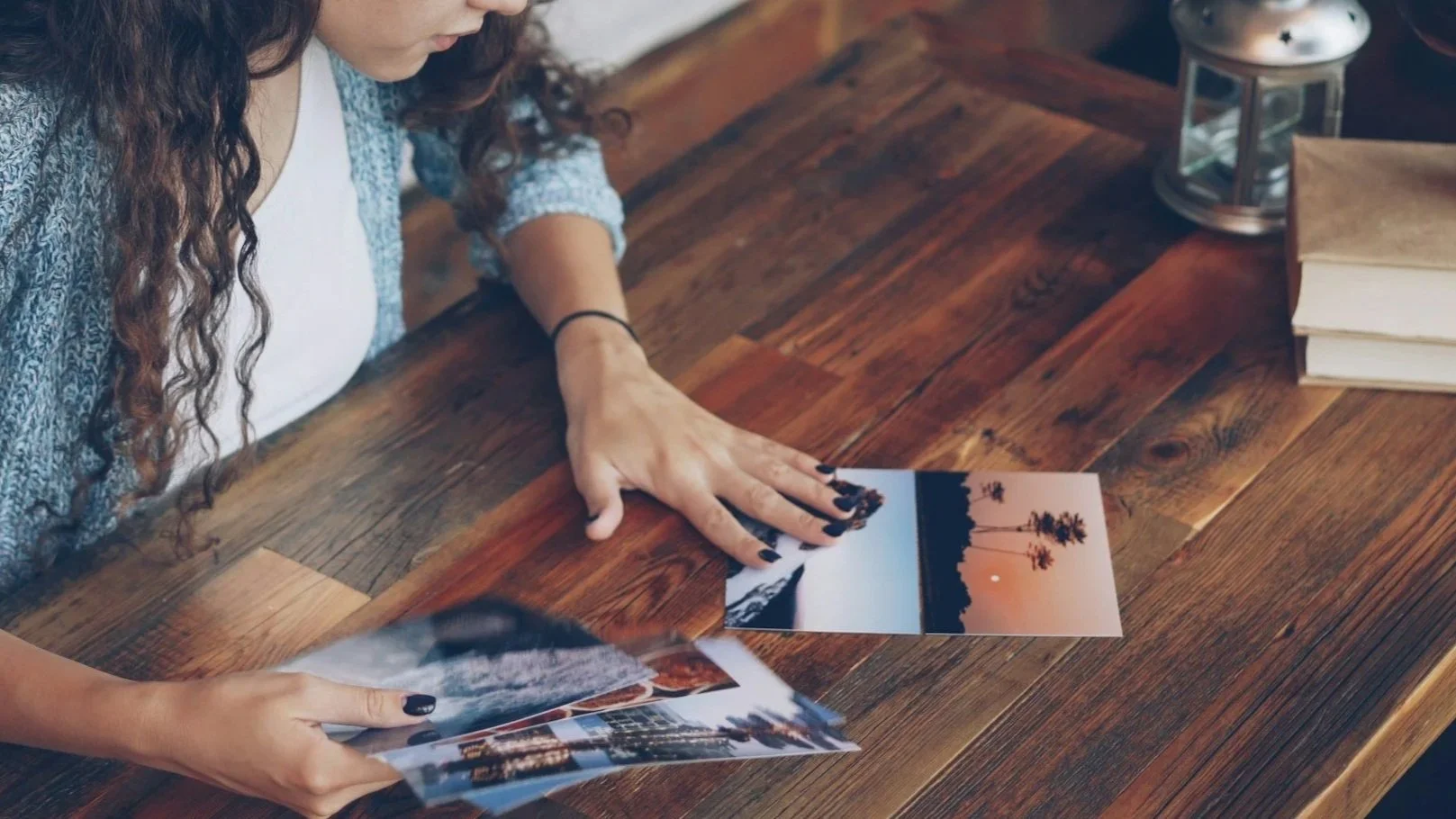 A woman with long curly hair is looking at and sorting through printed photographs on a wooden table.
