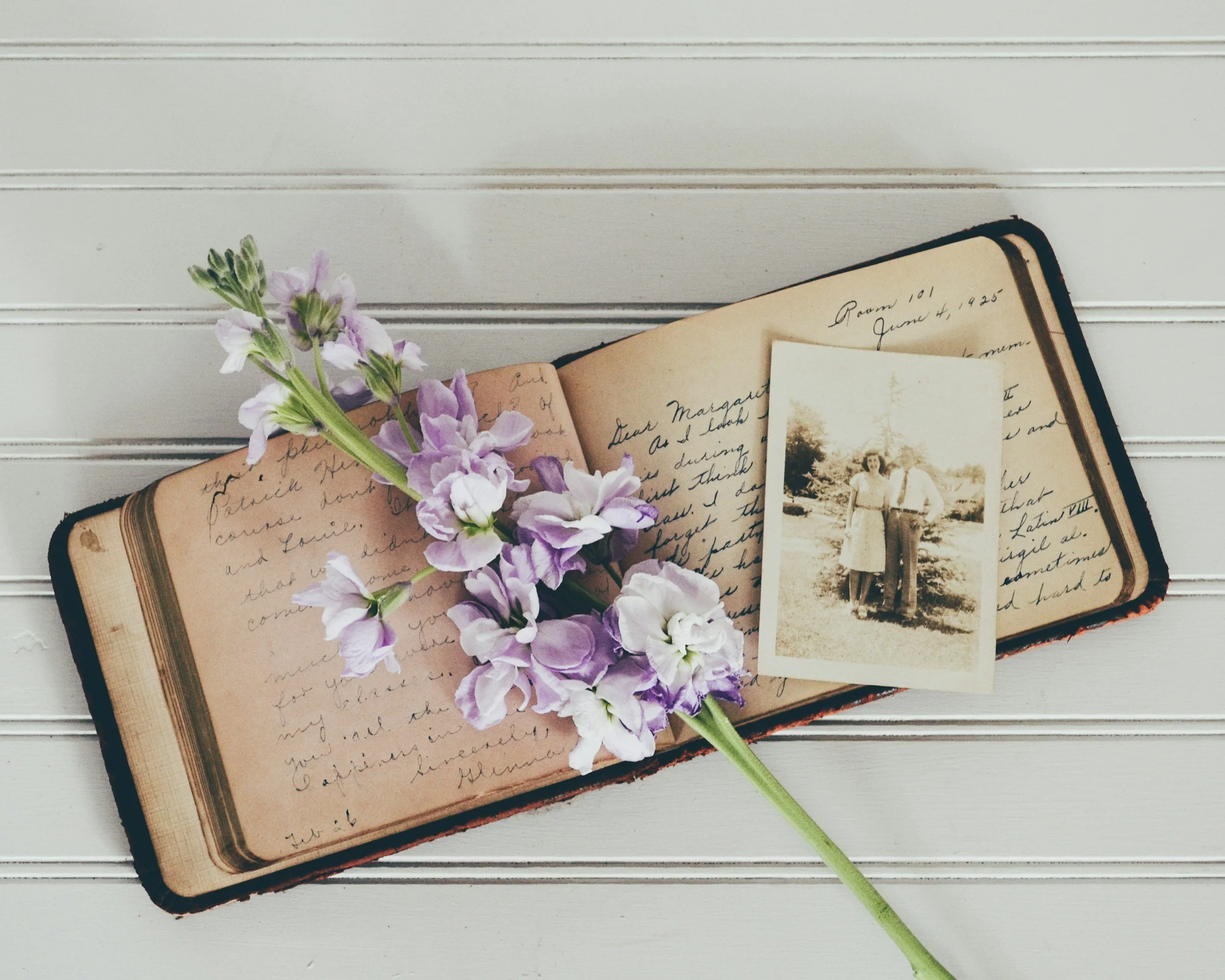 Open vintage journal with handwritten notes, black-and-white photograph of two women, and a stalk of purple and white flowers resting on the journal page, on white wooden surface.