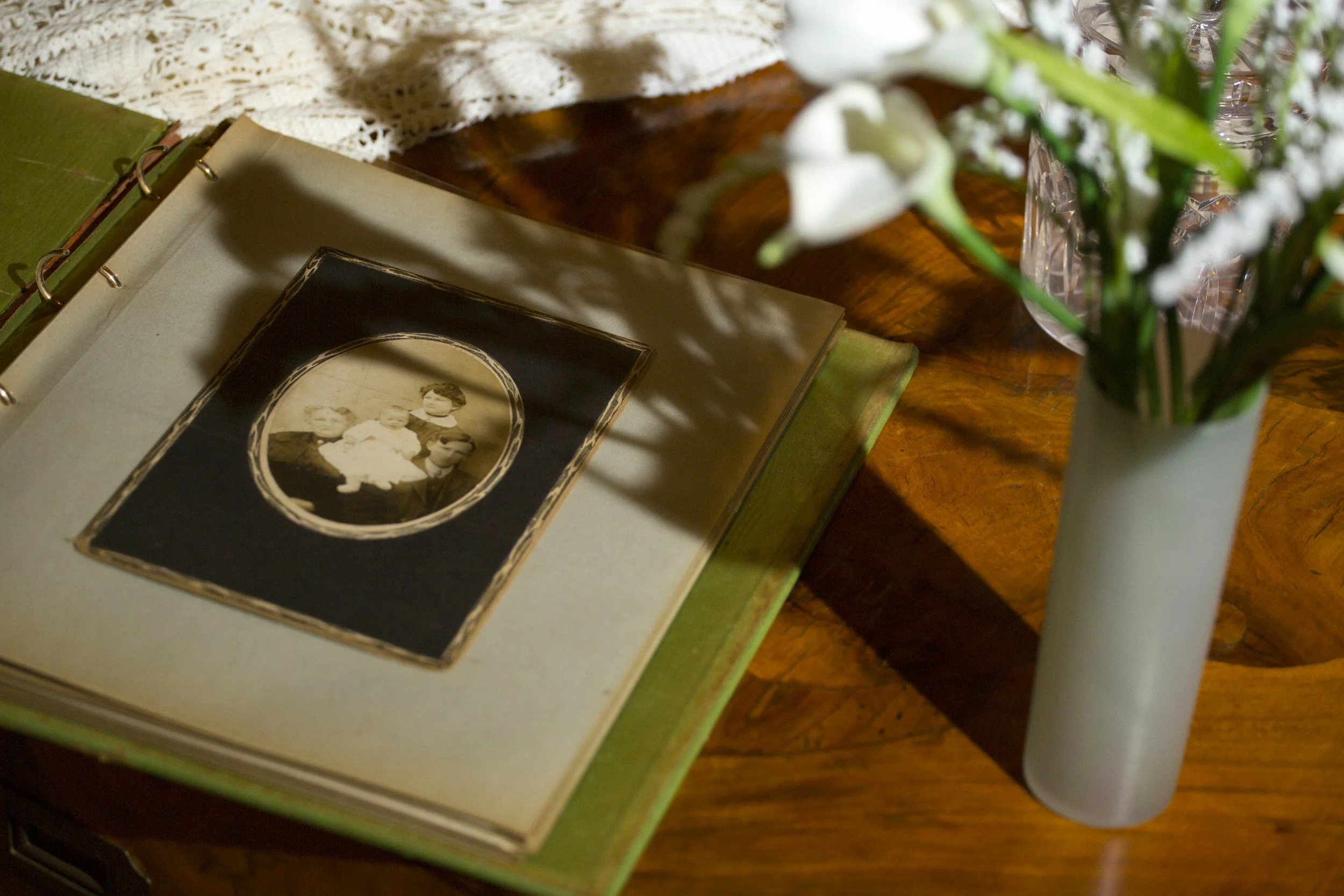 A table with a vintage photograph of three children, an open green photo album, a lace cloth, and a white vase with flowers.