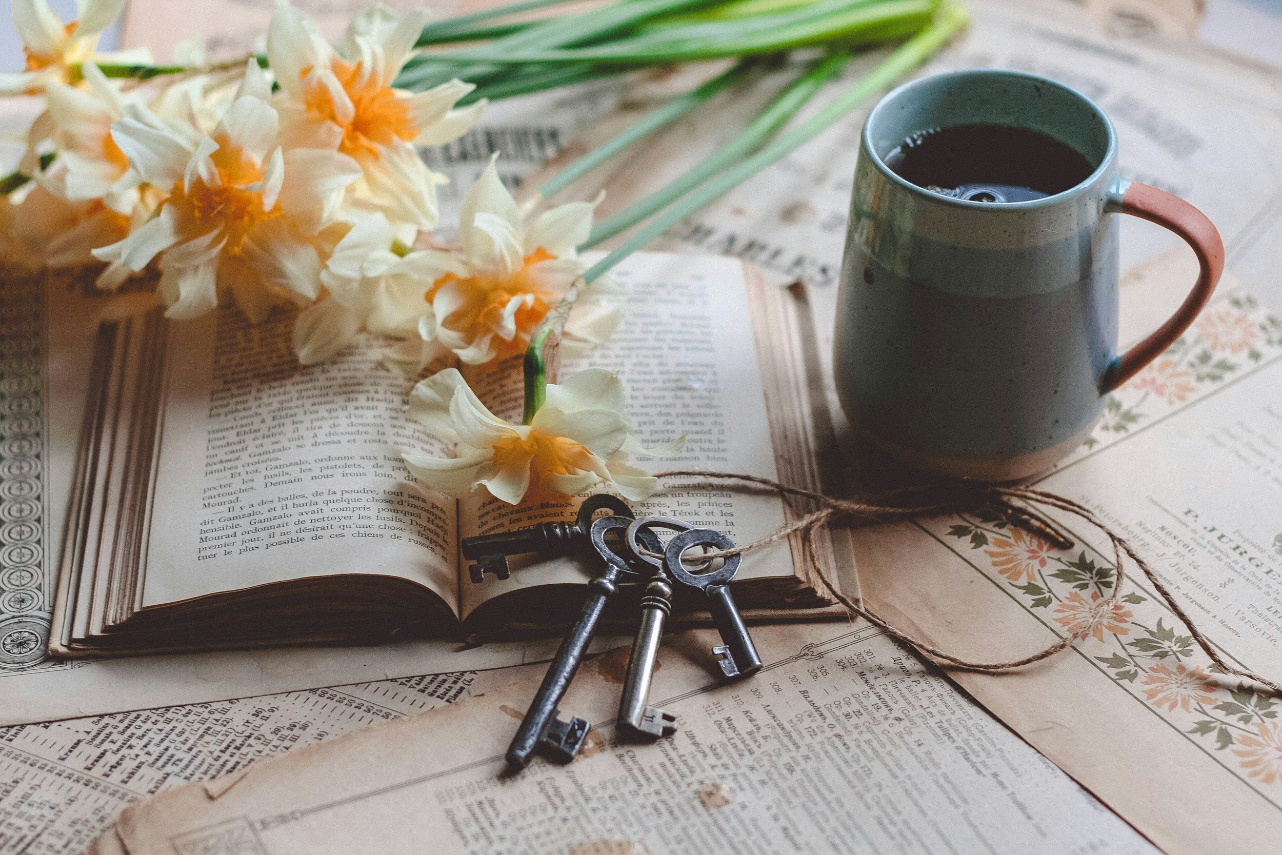 An open vintage book with flowers, keys, and a cup of coffee on a table covered with pages and newspapers.