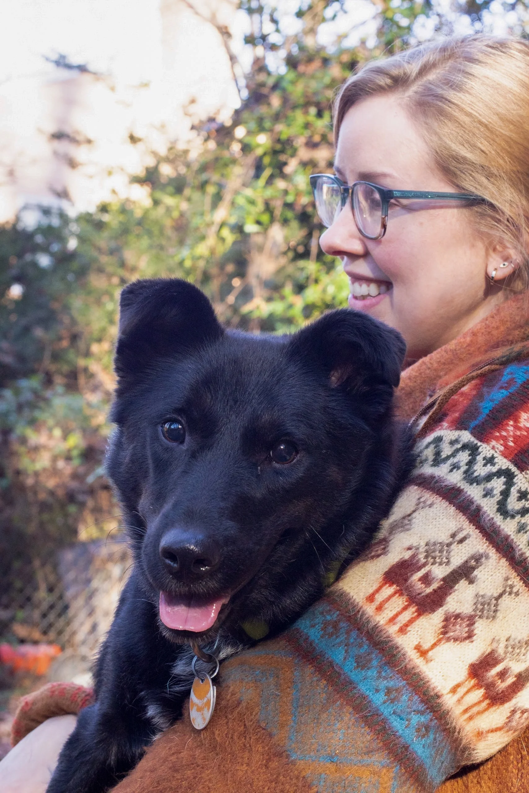 A young woman with glasses and earrings smiling, holding a black puppy with a collar and tag, outdoors with trees in the background.