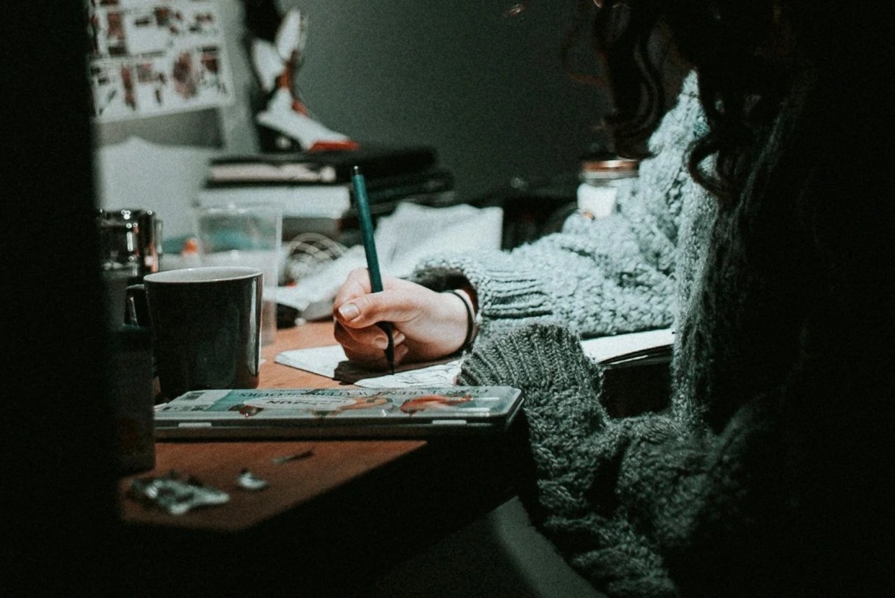 A person with long dark curly hair wearing a thick gray sweater, sitting at a cluttered table writing or drawing with a green pen on paper or a notebook.