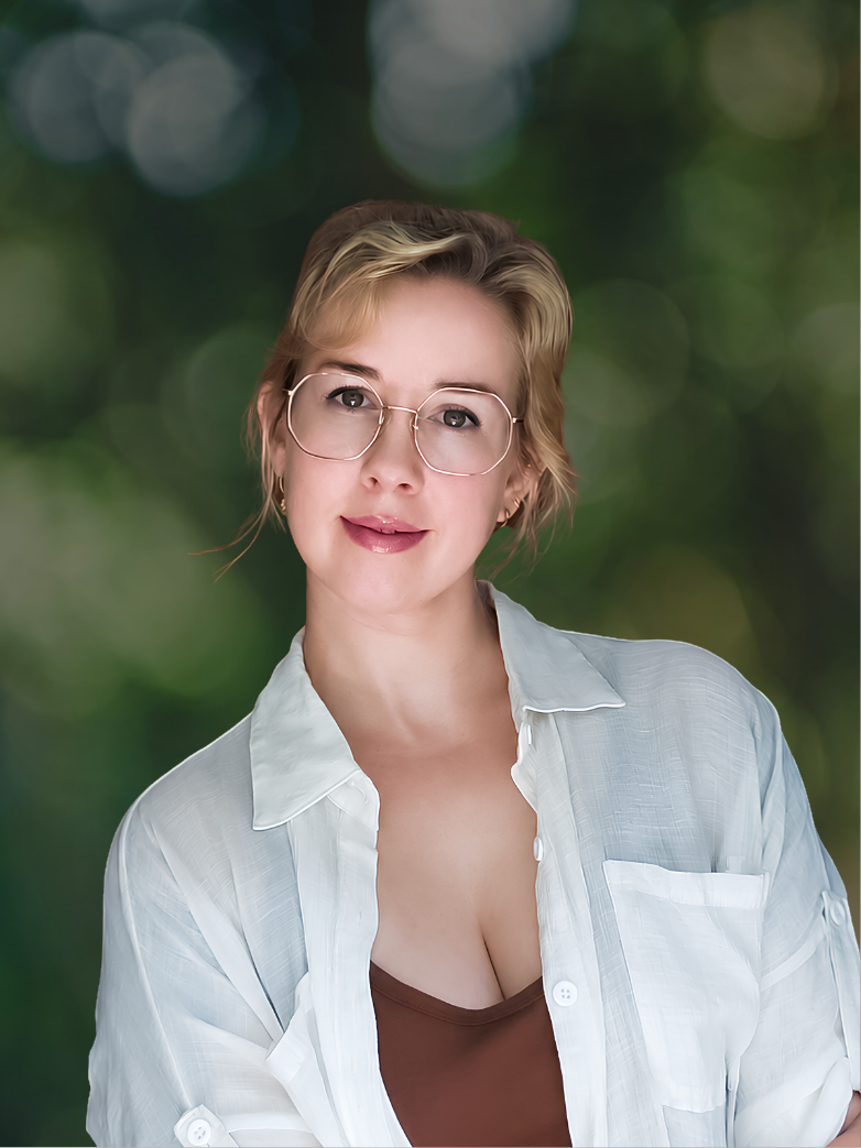 Portrait of a woman with short blonde hair, wearing glasses, a white button-up shirt over a brown top, with a blurred green background.