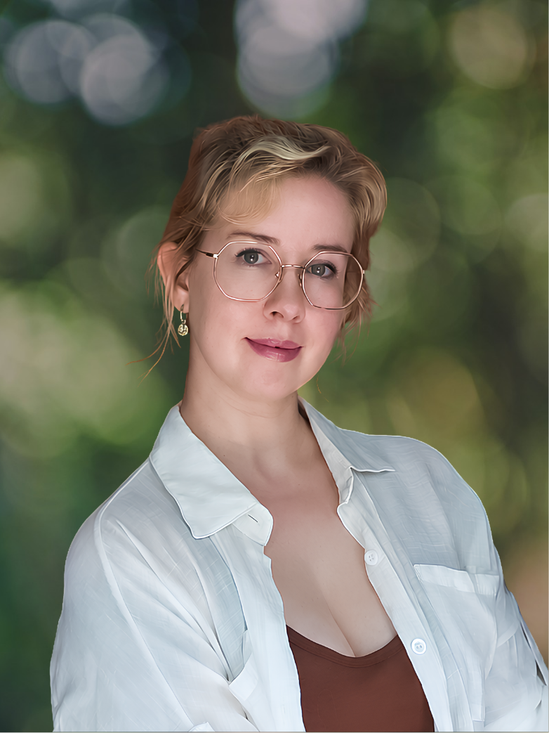A woman with short blonde hair, wearing glasses, a white shirt, and a brown top, standing outdoors with a blurred green natural background.