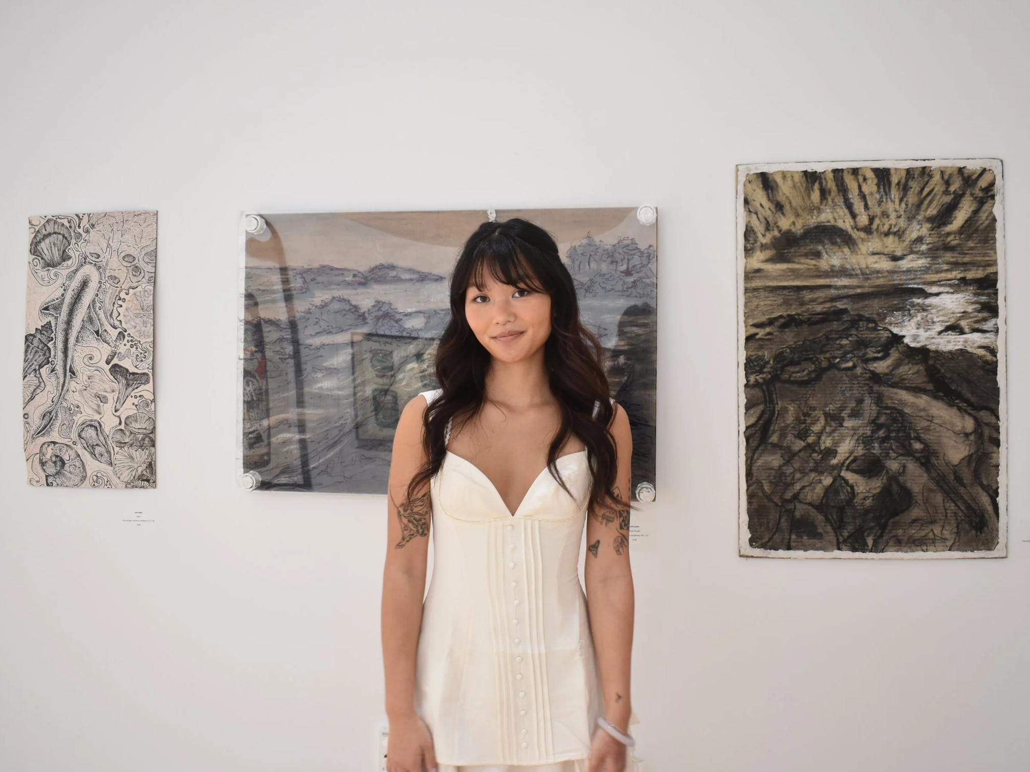 A woman with long dark hair and tattoos on her arms standing in front of framed artwork at an art gallery.