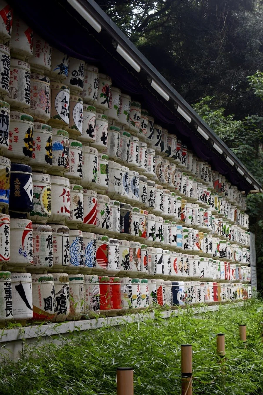 A wall lined with rows of traditional Japanese sake barrels with colorful artwork and Japanese writing, set outdoors with greenery and trees in the background.