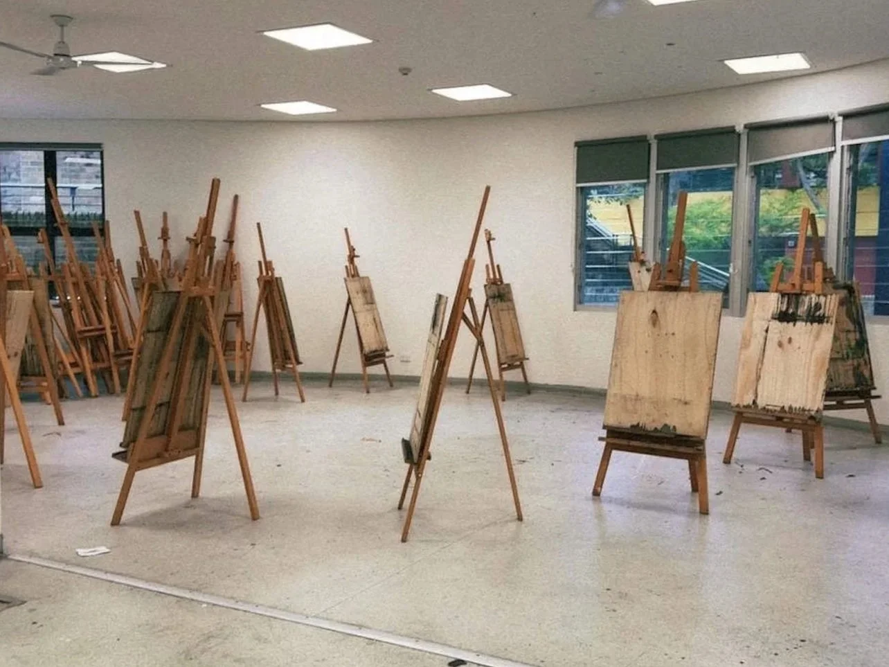 An art classroom with empty wooden easels and canvas boards arranged on the floor, windows on the right side, and a plain white wall in the background.