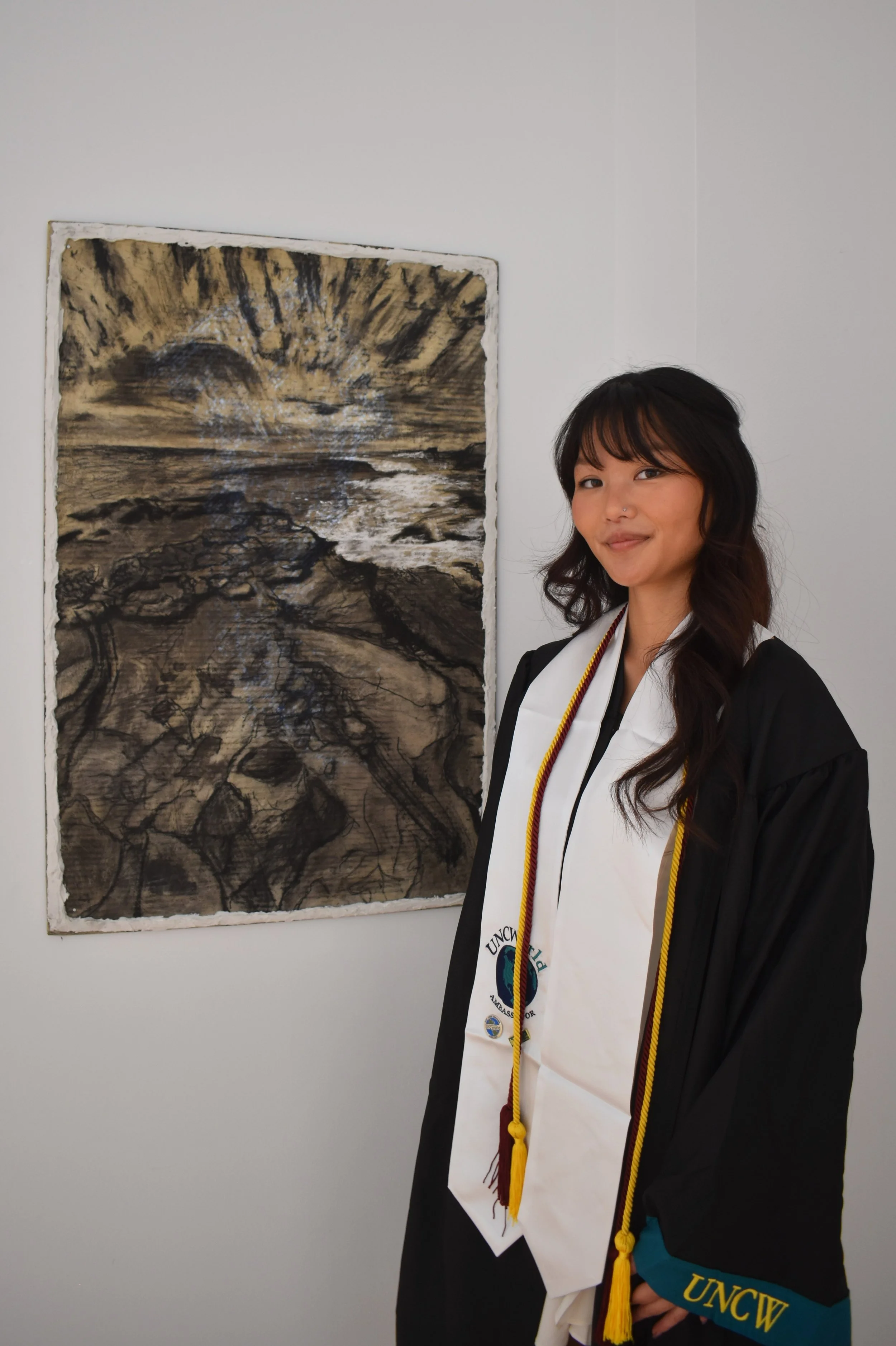 A woman in graduation attire standing next to a large black and white artwork that appears to be a painting of a rocky beach with waves and a cloudy sky.
