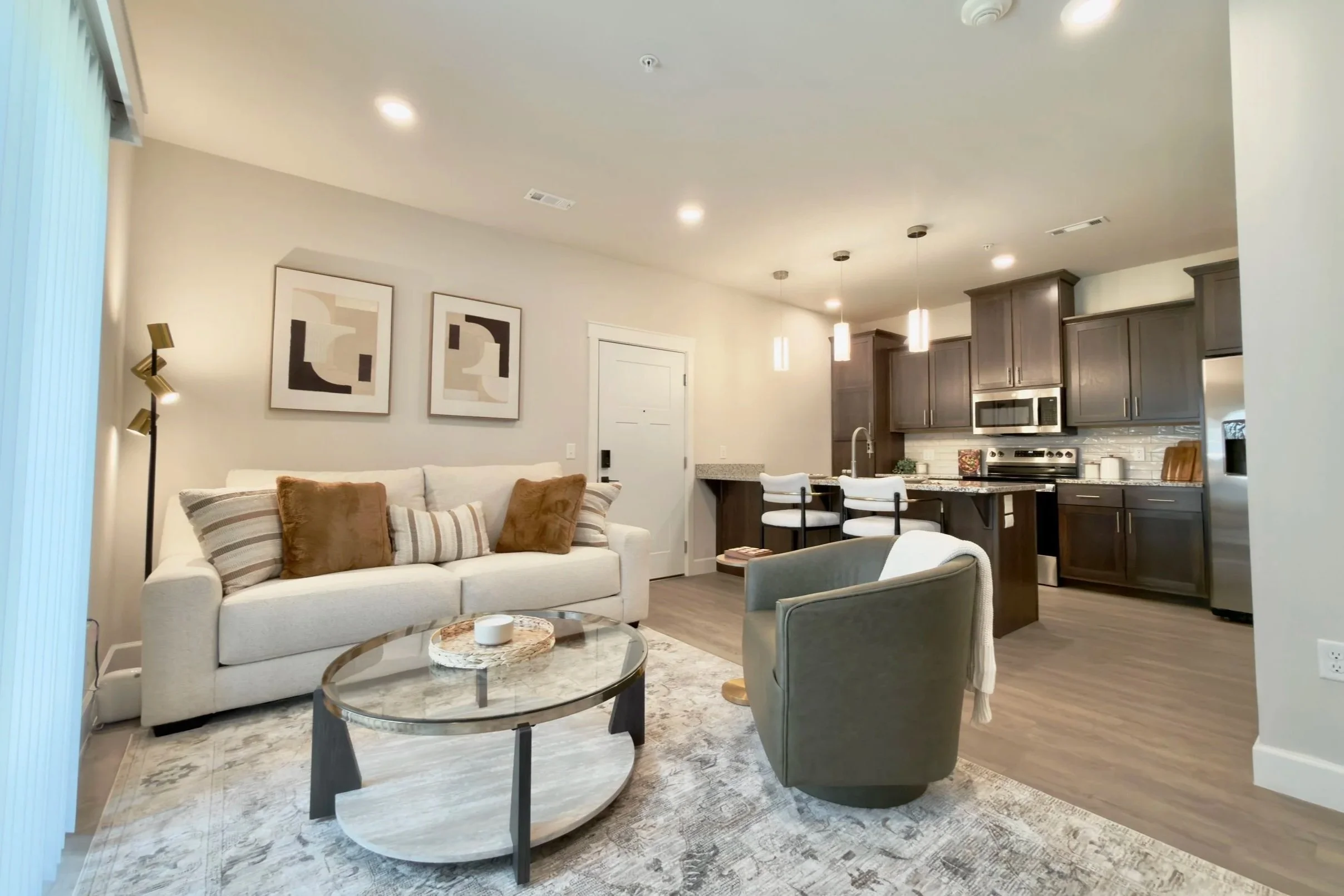 Living room with a cream sofa, striped pillows, a glass coffee table, two armchairs, art on the wall, and part of a kitchen with dark cabinets, a stainless steel microwave, and pendant lights.