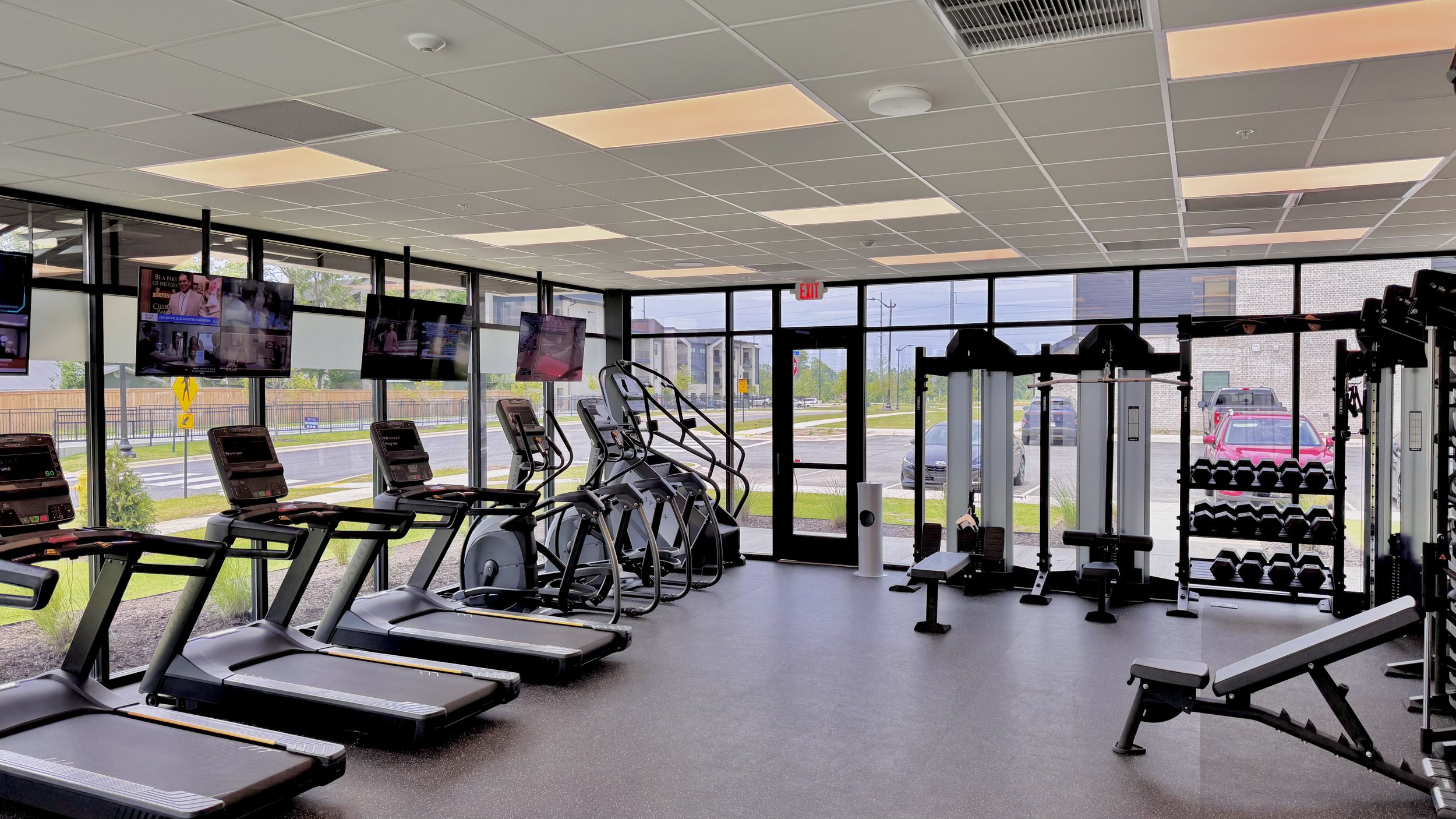 Interior of a gym with treadmills, ellipticals, weight machines, and TV screens, with large windows showing parked cars outside.