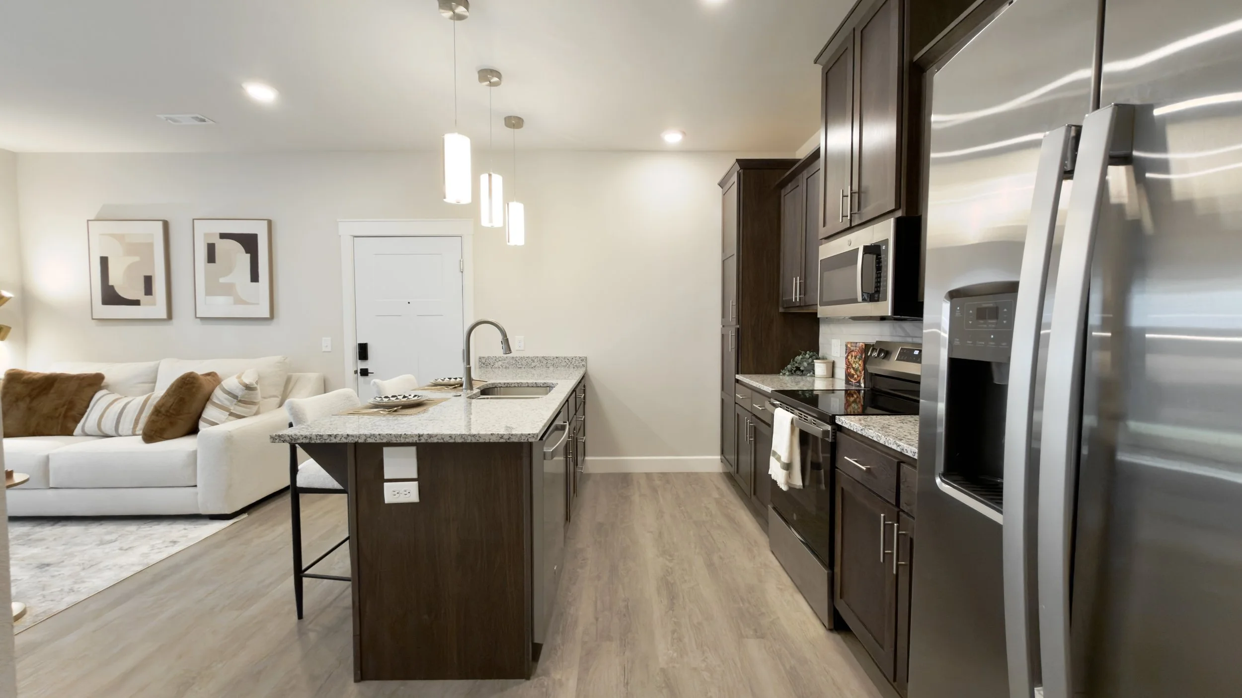 Modern kitchen with dark wood cabinets, stainless steel refrigerator, microwave, and oven, adjacent to a living room with a white sofa and abstract artwork on the wall.