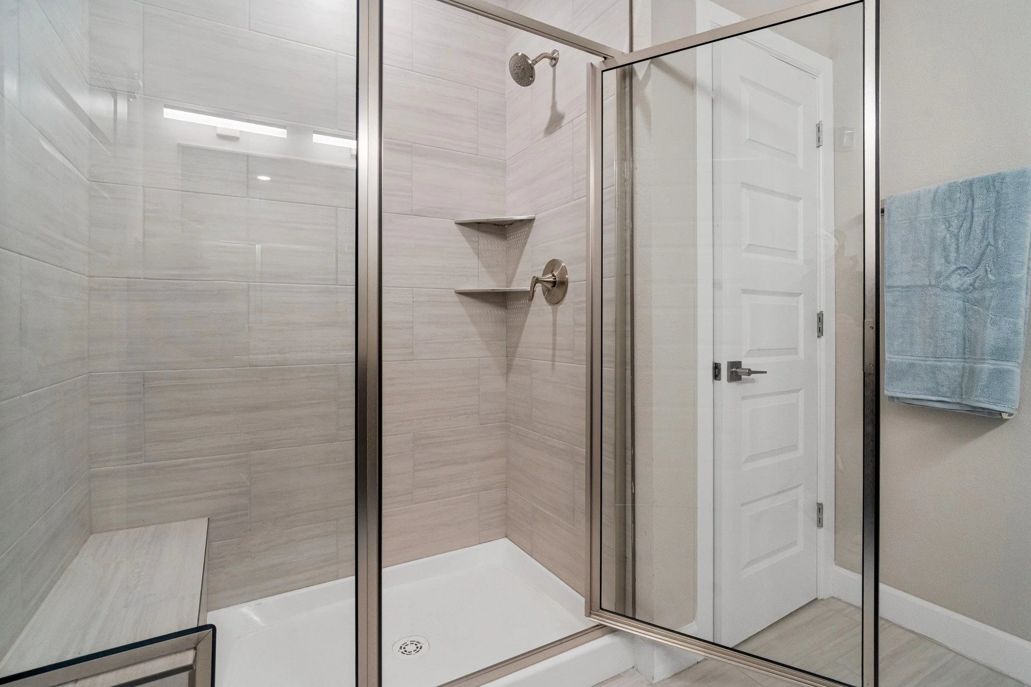A modern shower enclosure with clear glass walls and a white shower tray, featuring beige tiled walls, a showerhead, a faucet, and two corner shelves, next to a closed white door with a blue towel hanging on a towel rack.