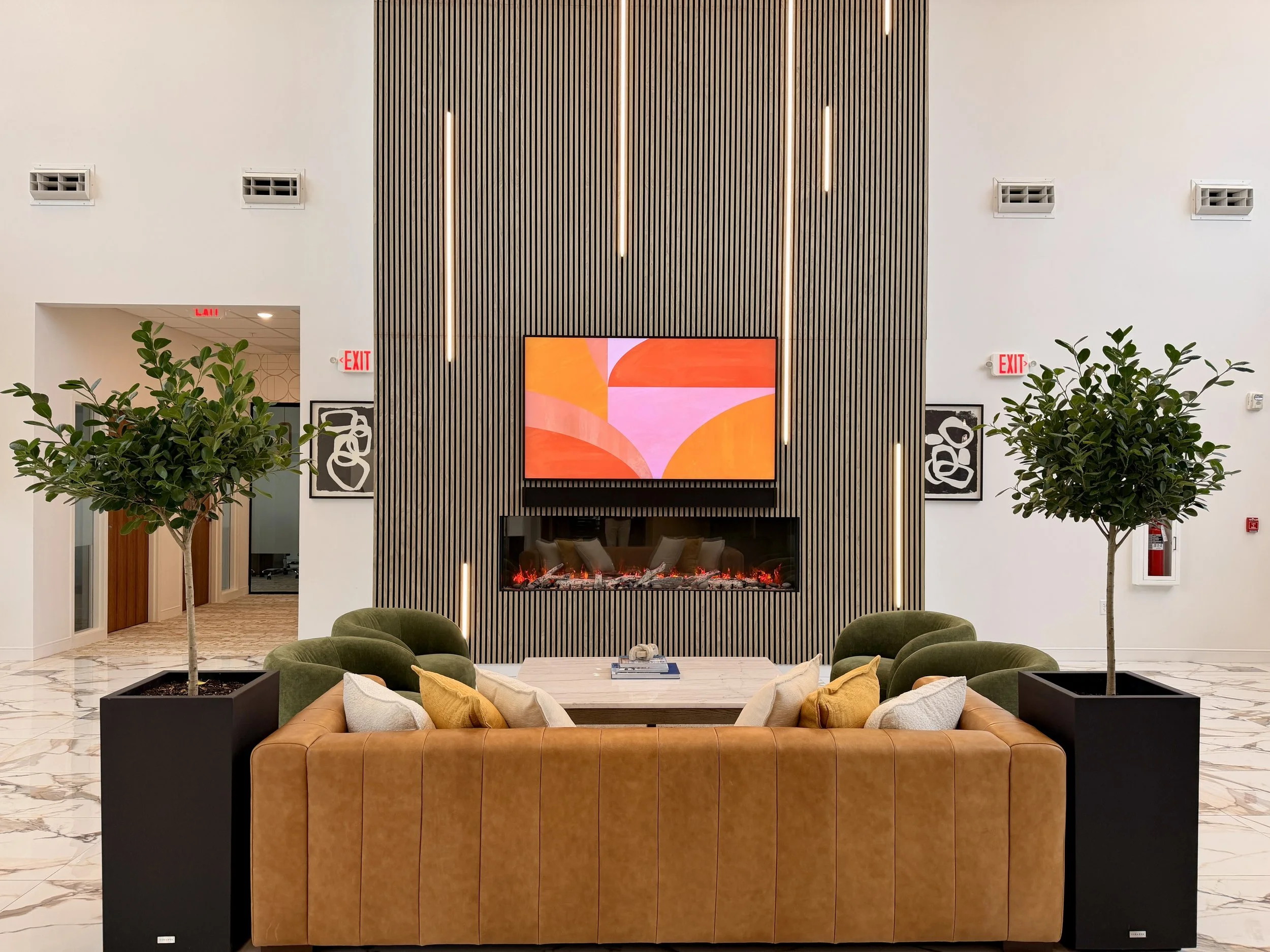 Interior of a modern lounge or waiting area featuring a marble floor, a brown leather sofa with yellow and white cushions, two black planters with leafy green plants, and a wall with vertical wooden slats. There are several framed artworks on the walls, a digital art display, and visible air vents, with a fireplace beneath the art display.