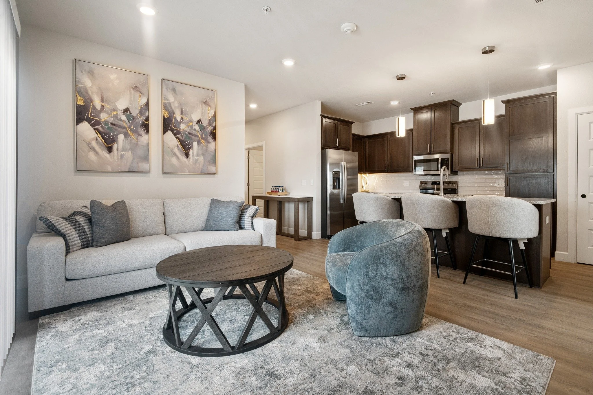 Living room with white sofa, abstract wall art, glass-top coffee table, armchair, and open kitchen with dark wood cabinets and stainless steel appliances.