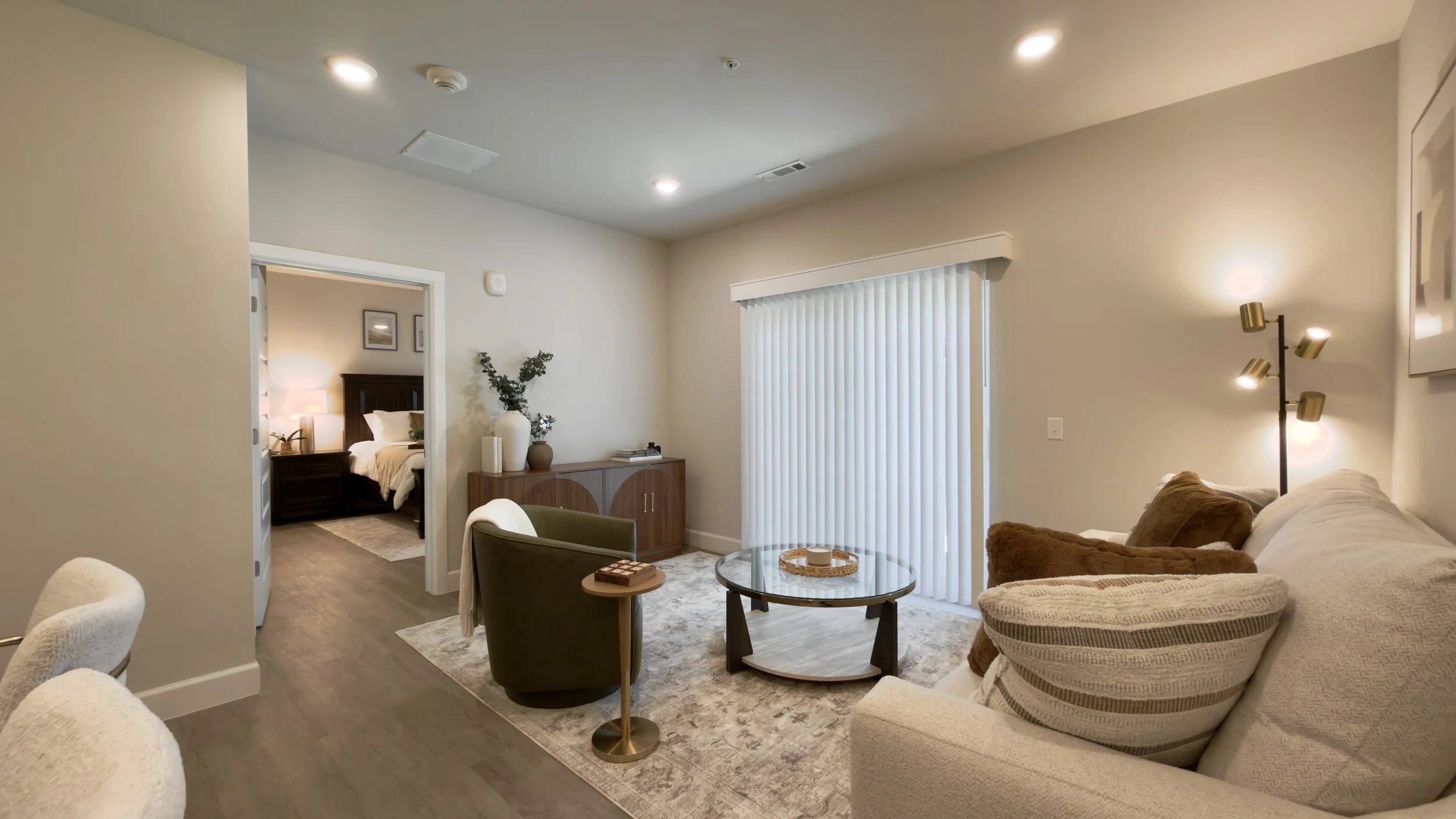 Living room with beige walls, a large window with vertical blinds, a beige sofa with pillows, a glass coffee table, a black armchair, a sideboard with decorative vases and plants, and a floor lamp with multiple lights. In the background, a bedroom wi