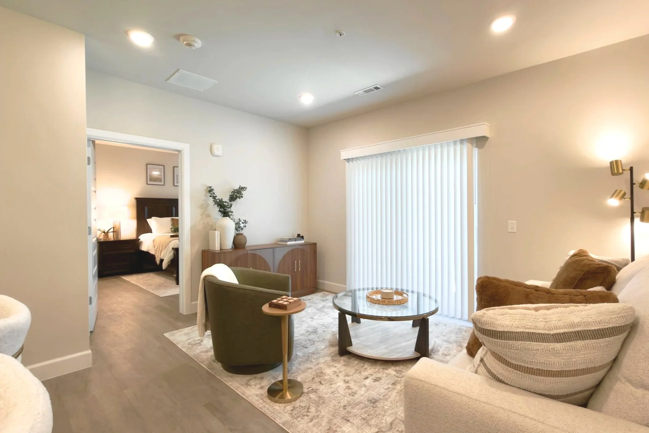 A living room with beige walls, a glass coffee table, a beige couch with pillows, and an armchair. There is a wooden sideboard with decorative vases and a plant. A large sliding glass door with vertical blinds opens to the outside. A lamp with two ad