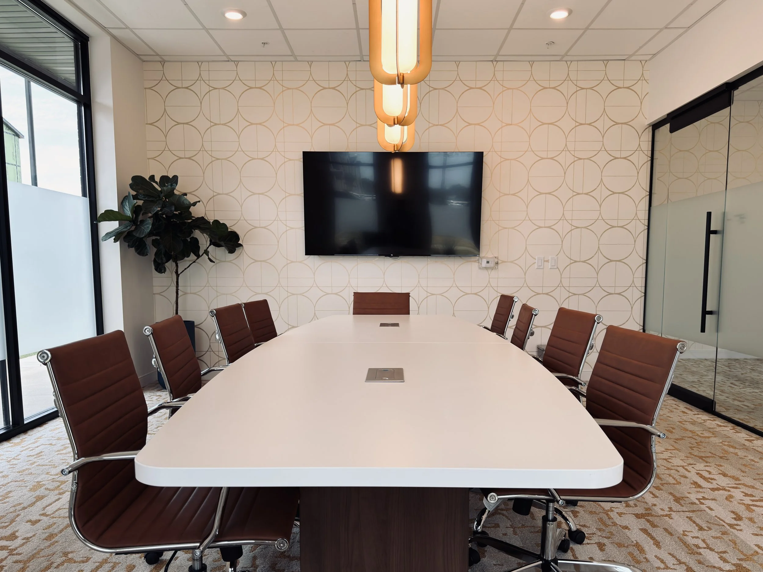 A modern conference room with a large white oval table surrounded by brown leather chairs. There is a wall-mounted flat-screen TV, a large window letting in natural light, and stylish lighting fixtures hanging from the ceiling. A potted plant is near the window, and the walls have a decorative geometric pattern.