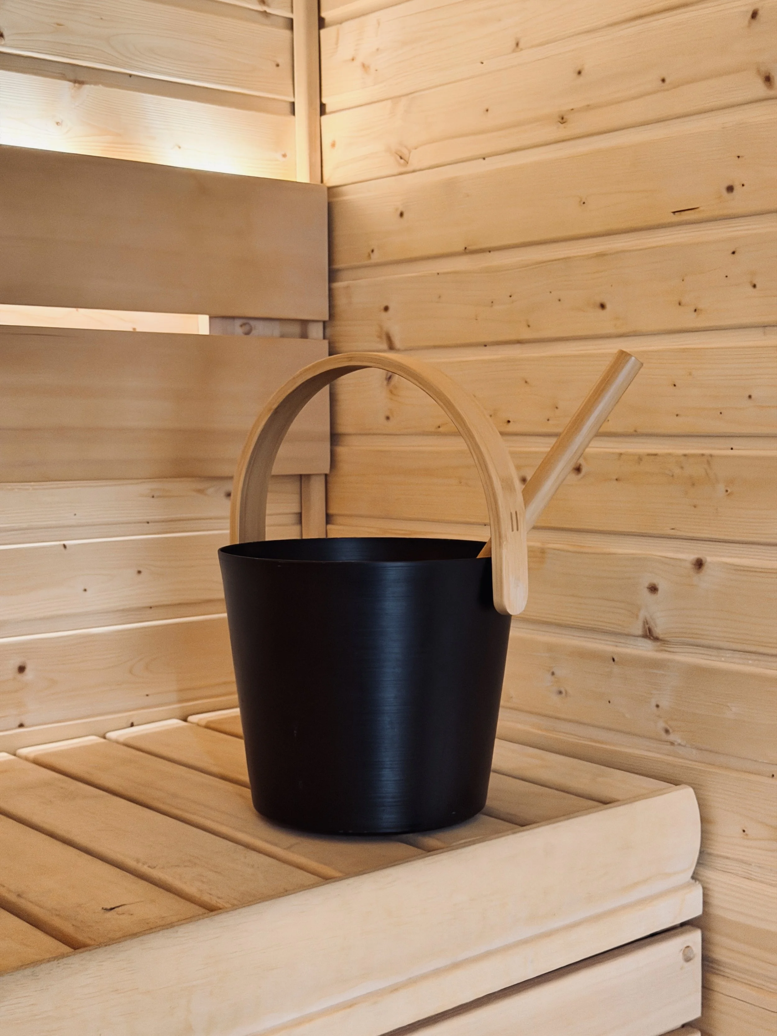 A black bucket with a wooden handle and a wooden straw is placed on a wooden shelf inside a wooden sauna.