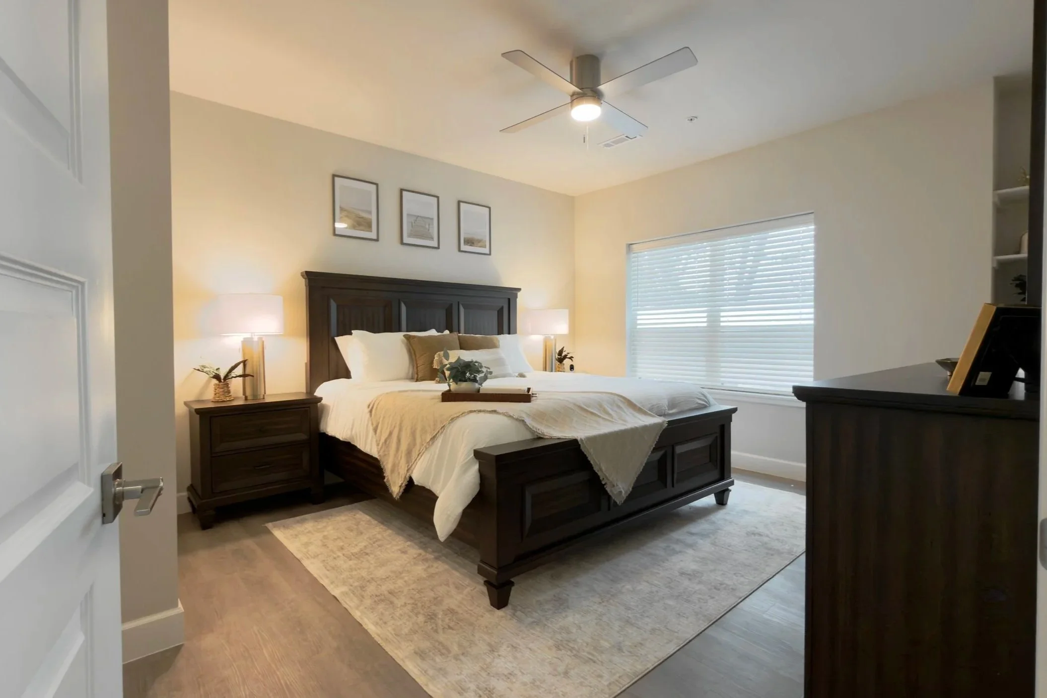 A bedroom with a dark wood bed frame, white bedding, and beige throw blankets. Two bedside lamps with white shades and plants sit on matching dark wood nightstands. The walls are light-colored, with three framed pictures hanging above the bed. A larg