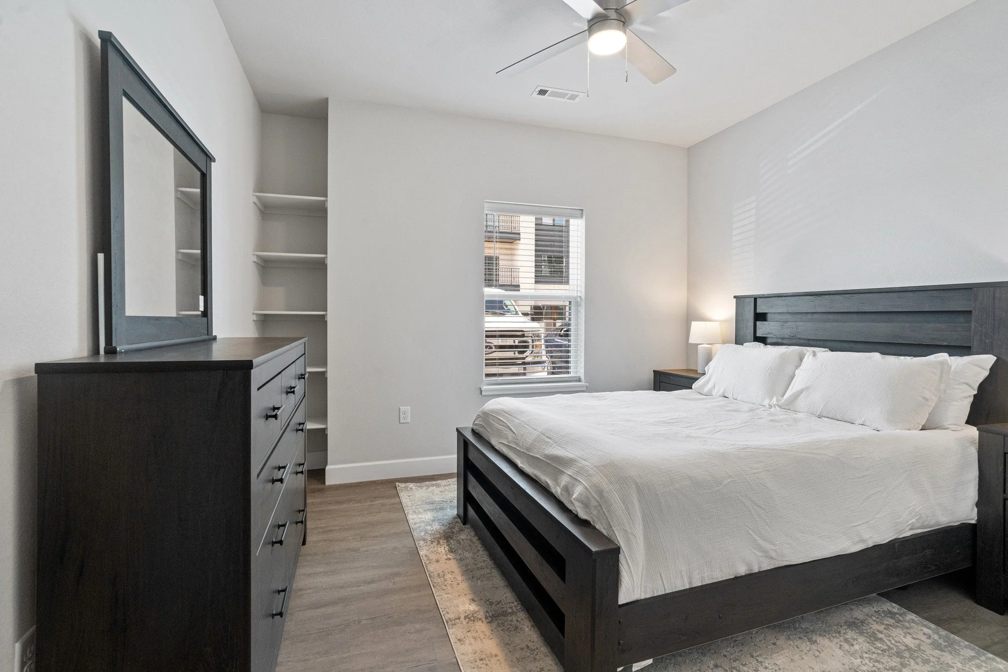A bedroom with a large bed, dark wooden frame, and white linens. There is a dark wooden dresser with a mirror, a nightstand with a lamp, and a window with blinds.