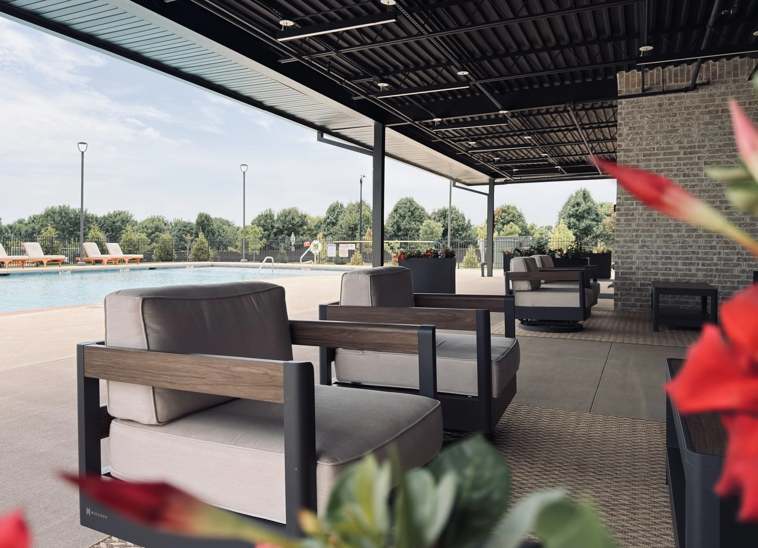 Outdoor swimming pool area with lounge chairs, a shaded sitting area with chairs and tables, surrounded by trees and a fence, under a partly cloudy sky.