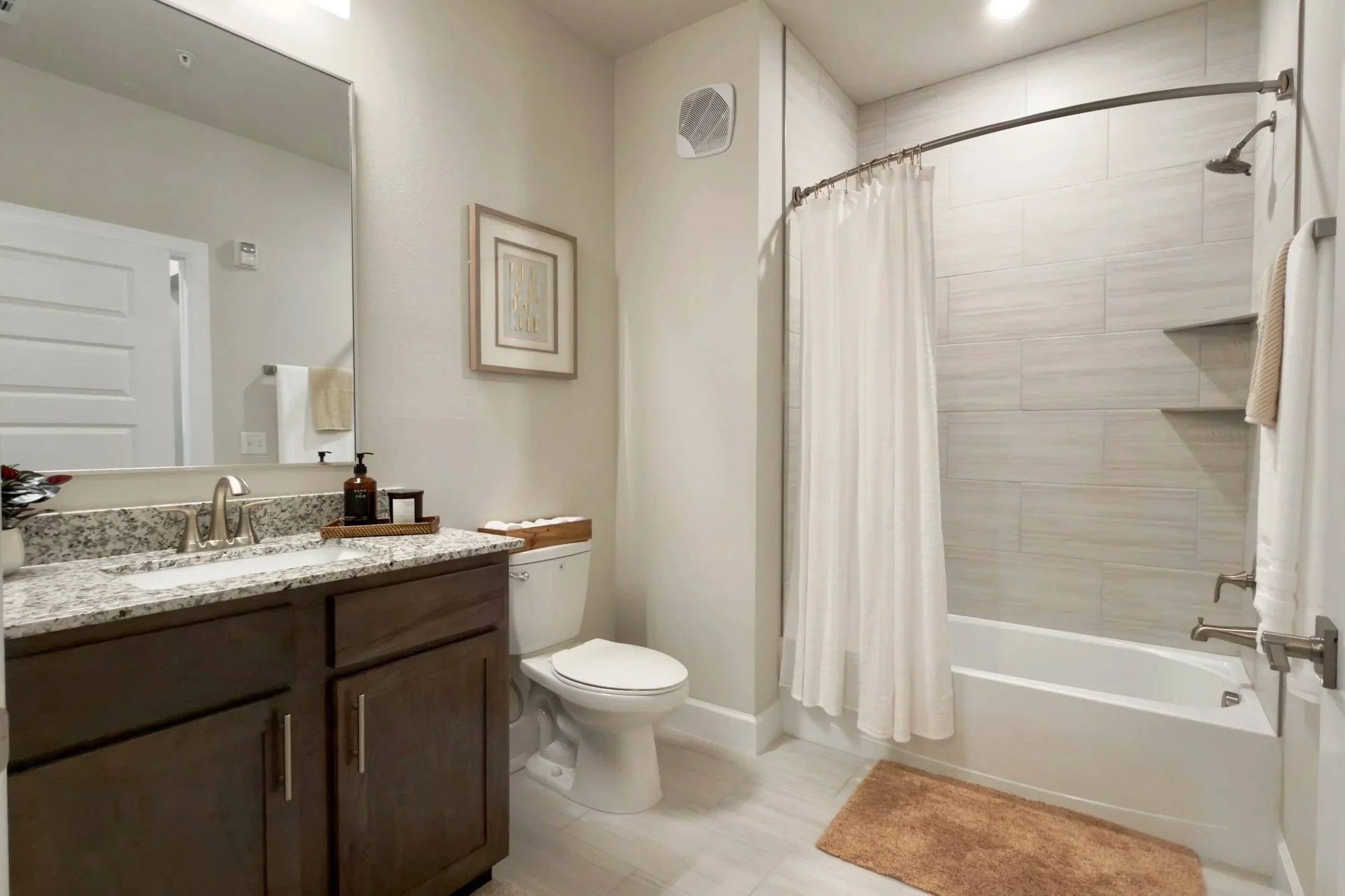 Bathroom with a granite countertop vanity, mirror, white toilet, shower area with a curtain, light-colored wall tiles, framed wall art, and a brown rug.