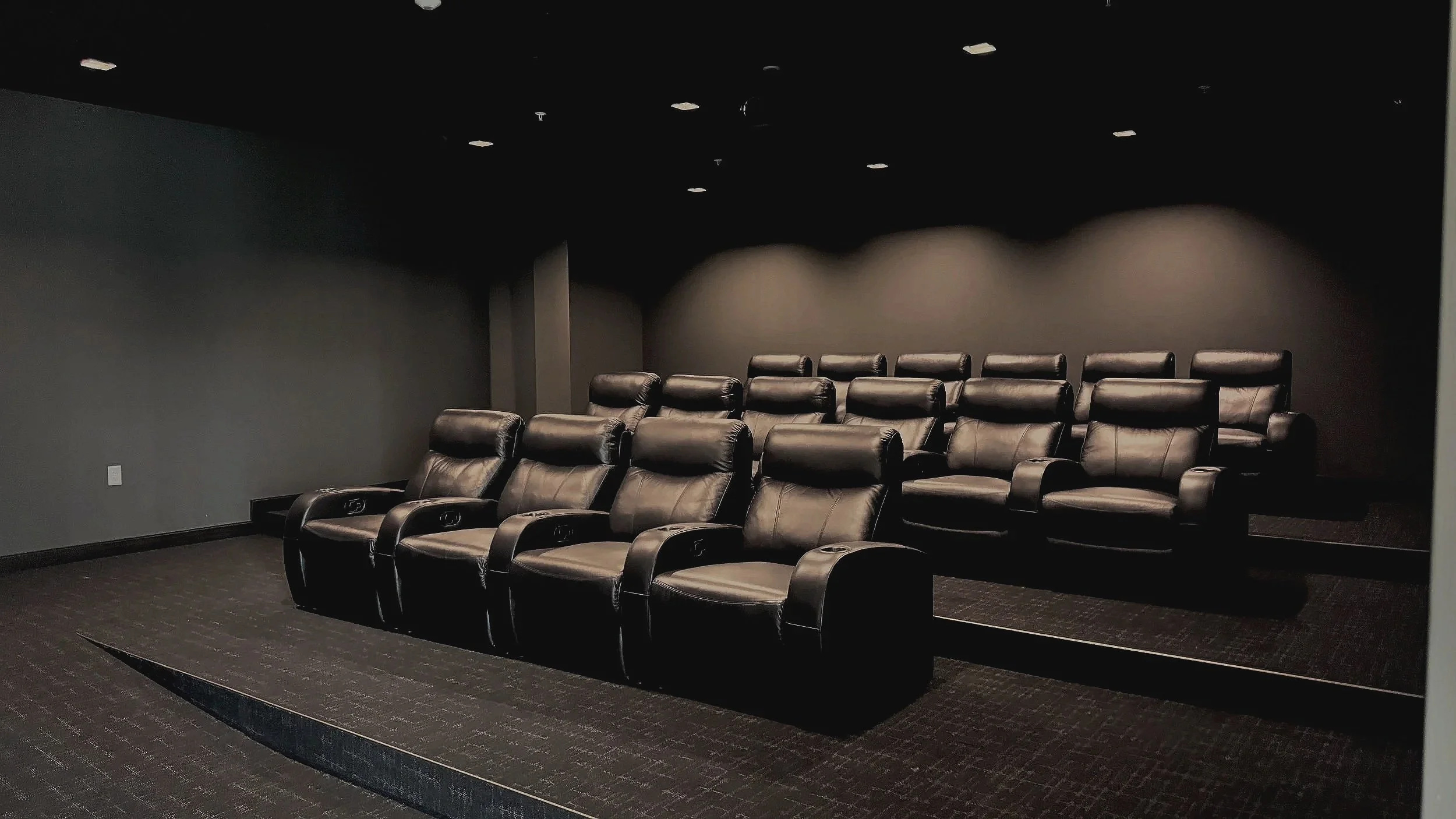 A small theater or screening room with two rows of black leather recliner chairs on a raised platform, dark walls, and ceiling lights.