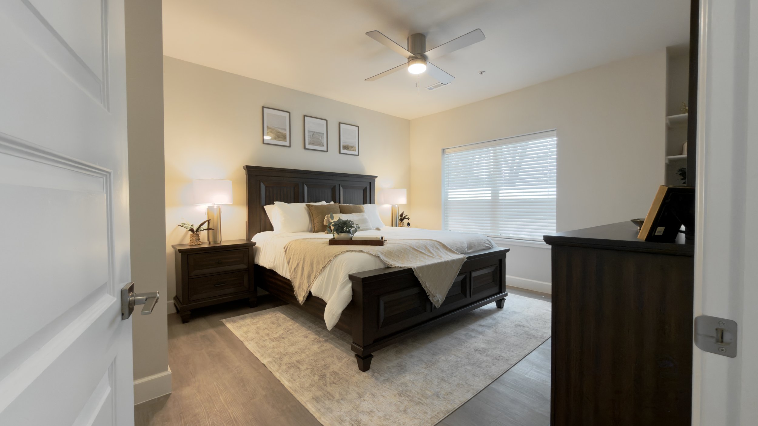 A bedroom with a wooden bed frame, beige bedding, two side tables with lamps, and framed artwork on the wall. There is a window with blinds and a ceiling fan with a light.