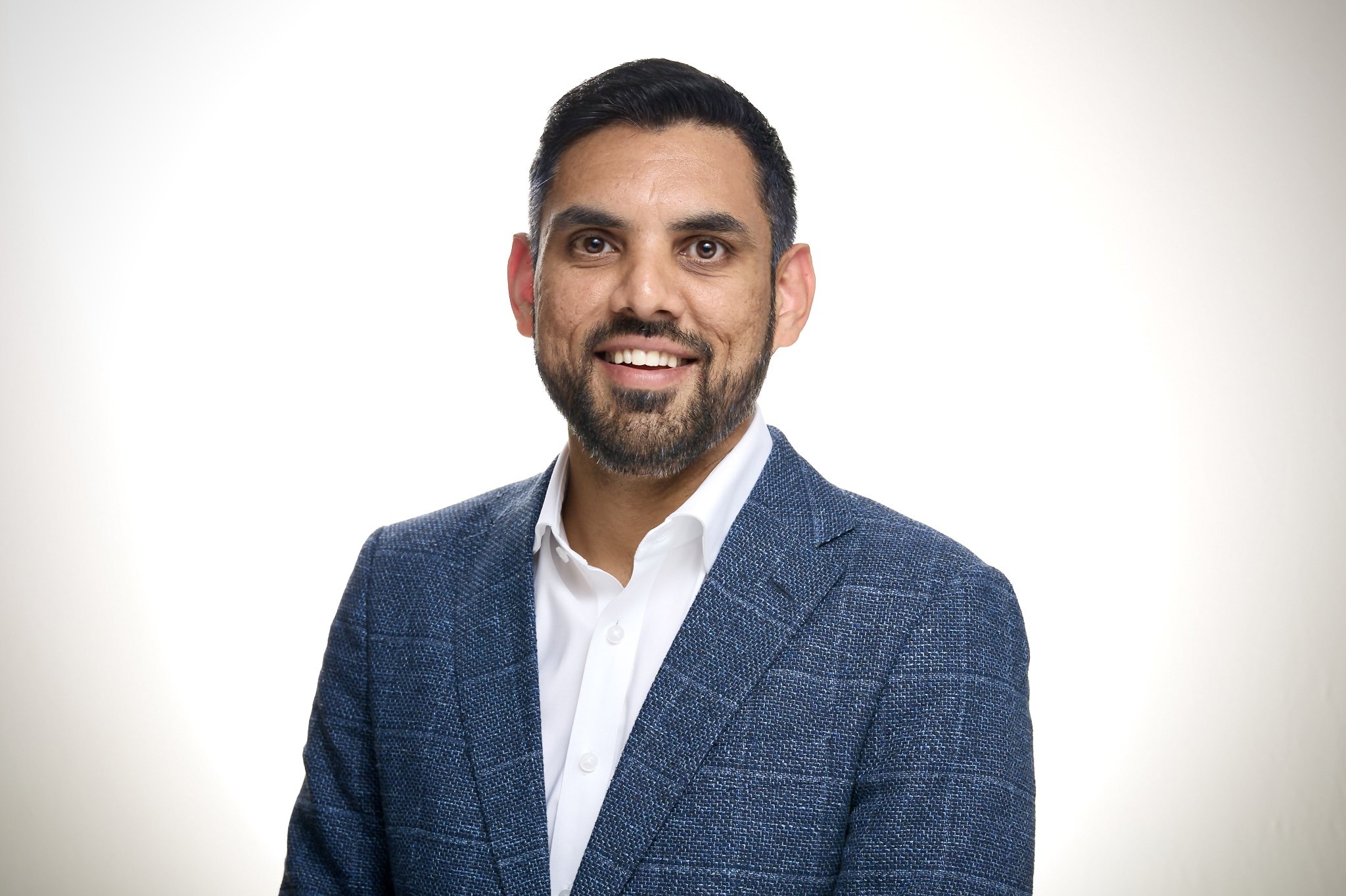A man in a blue checked blazer and white shirt smiling at the camera against a plain white background.