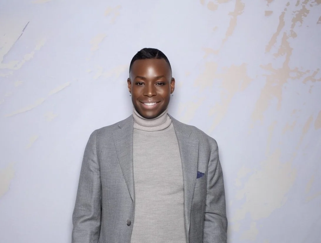 A person with dark skin, styled hair, wearing a gray blazer over a light gray turtleneck sweater, standing against a lightly textured light background and smiling.