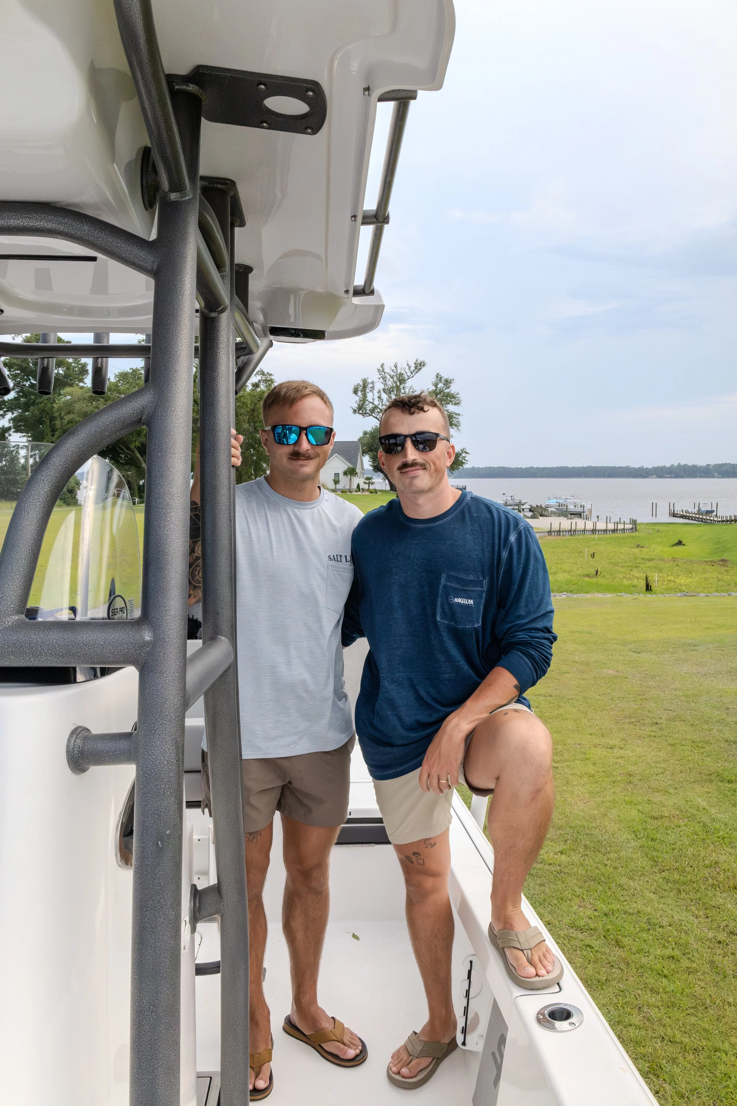 Two men standing on the deck of a boat by a lake, wearing sunglasses, casual clothing, and flip-flops, with a backdrop of water, docks, and grassy land.