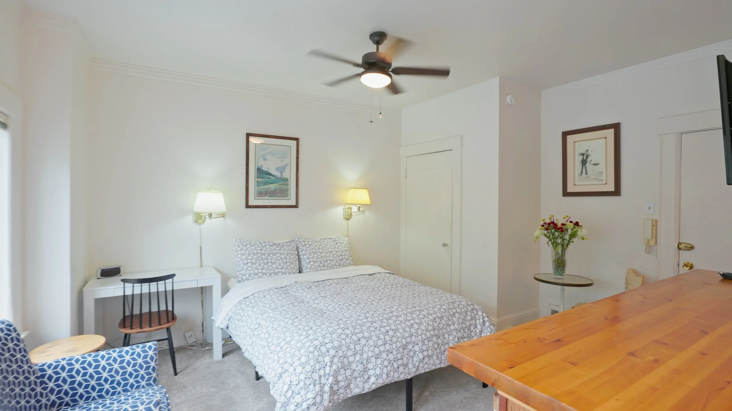 Woodland Ave Studio showing well-lit room with bed, arm chair, desk, cafe table, closet door, kitchen counter and front door.