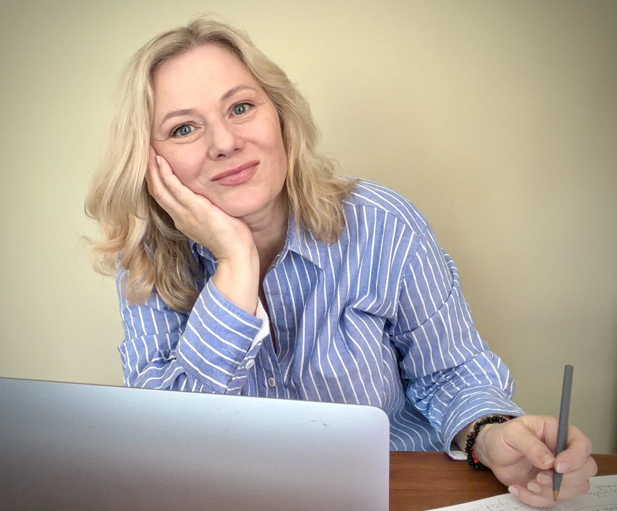 A blonde woman wearing a blue and white striped shirt is sitting at a table, resting her chin on her hand, and smiling gently at the camera.