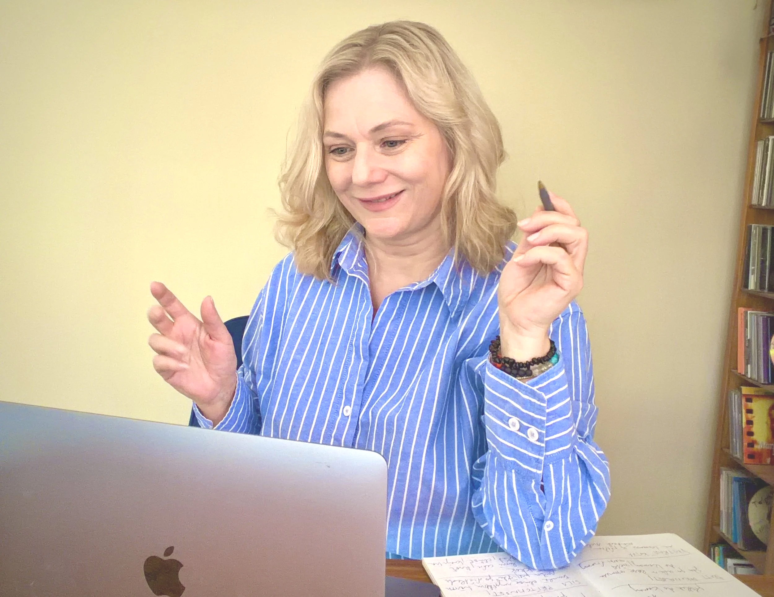 A woman with blonde hair wearing a blue striped shirt is sitting at a desk with a laptop in front of her. She is holding a black marker in her right hand and gesturing with her left hand. There is a piece of paper with handwritten notes on it on the desk. In the background
