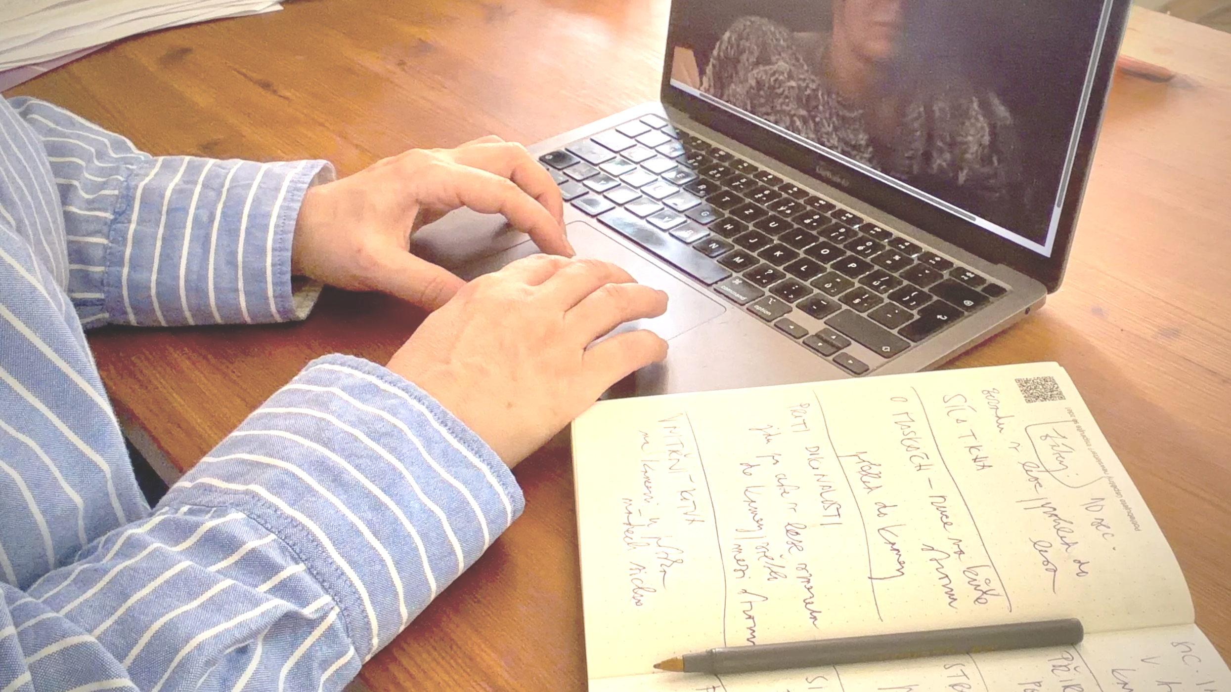 Person wearing a blue and white striped shirt writing notes in a notebook with handwritten text, a pencil, and a laptop on a wooden desk.