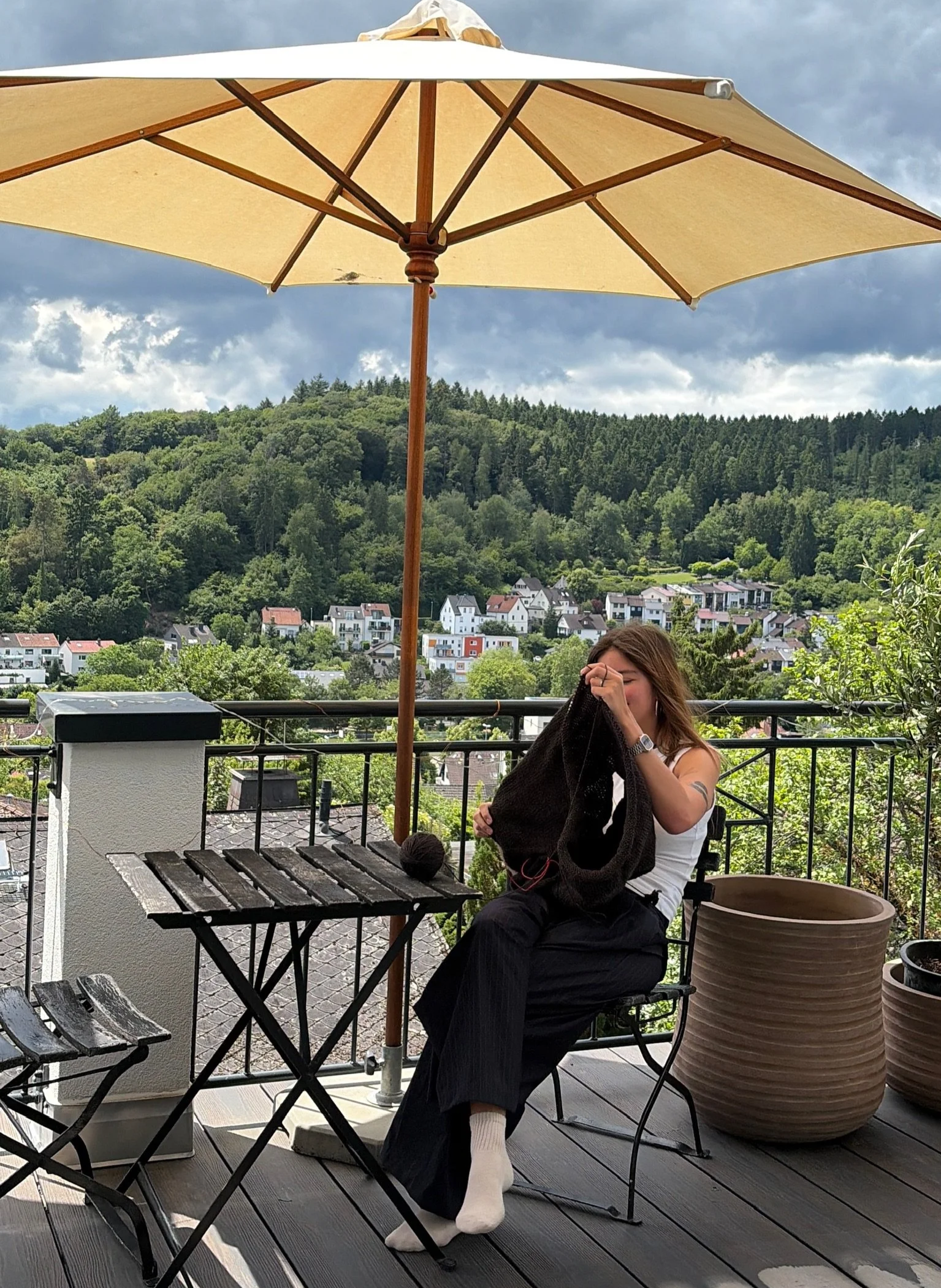 Eine Frau sitzt auf einer Terrasse mit Holzboden, umgeben von Töpfen und Gartenmöbeln. Sie trägt eine weiße Bluse, schwarze Hose, weiße Socken und strickt, während sie an einem schwarzen Kleid arbeitet.