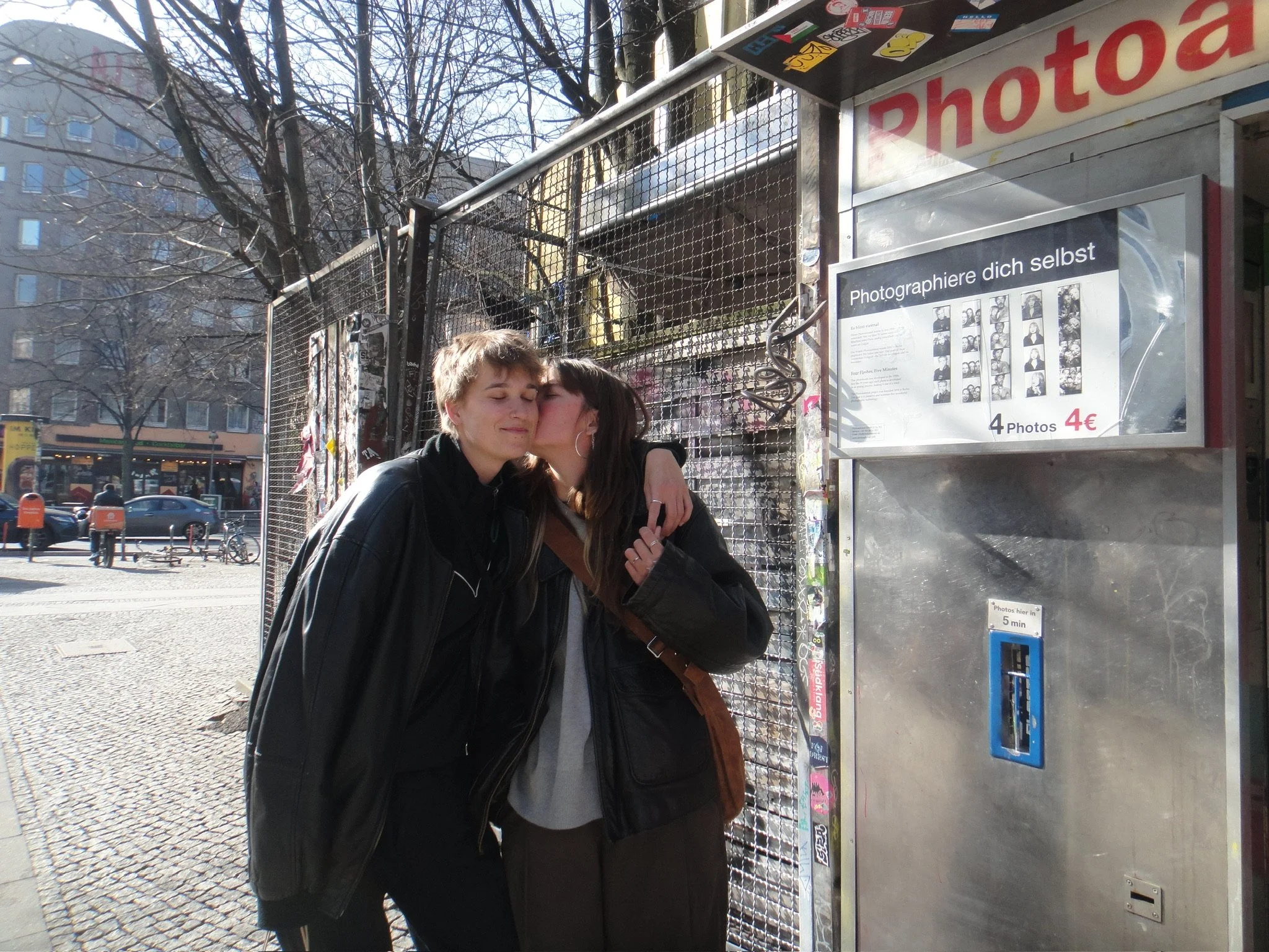 Zwei junge Frauen umarmen sich und küssen sich am Kopf vor einem Fotoautomaten im Freien, in einer belebten Stadt, bei sonnigem Wetter.