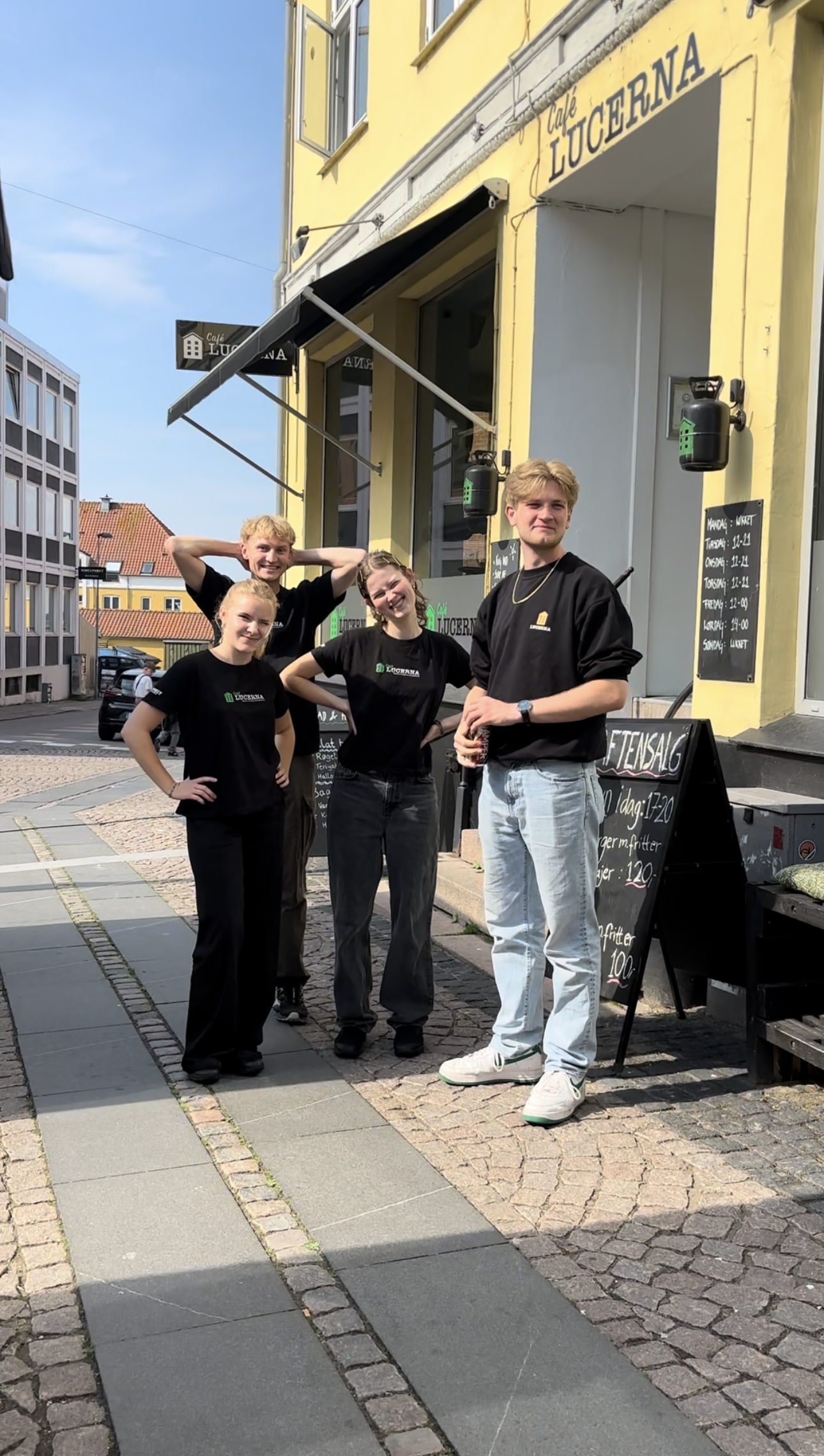 Fem unge mennesker udenfor en cafe, smilende og poserende for fotografiet, foran en gul bygning med cafe skilte og menu tavle.