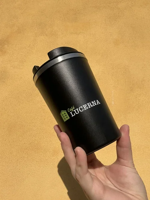 En hånd holder en sort termokande med logo for Café Lucerna mod en gul baggrund.