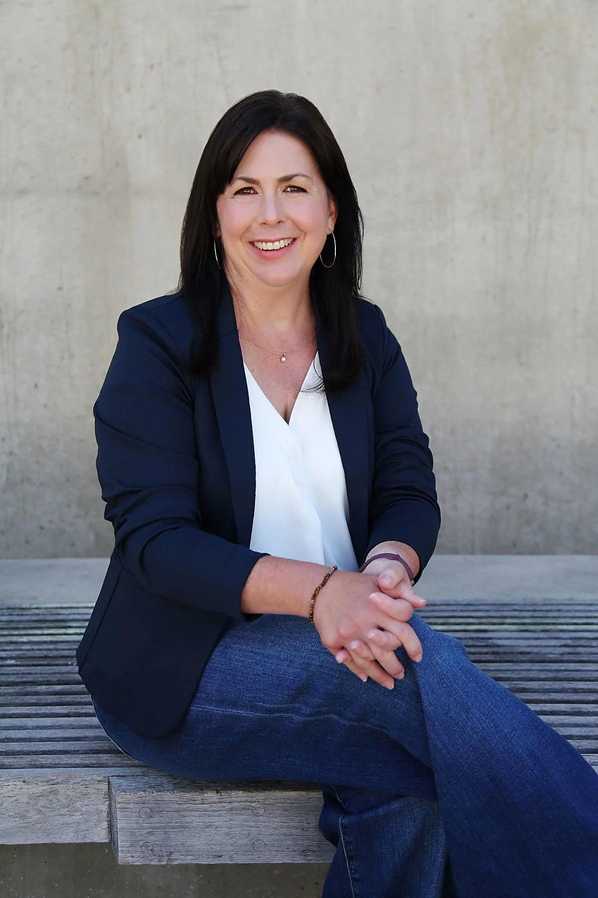 Kim Ocumen, leadership coach, wearing a navy blazer, white blouse, and jewelry, sitting outdoors on a wooden bench against a concrete wall, smiling at the camera.