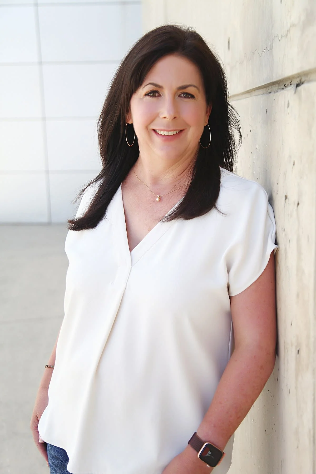 Kim Ocumen, leadership coach, with dark hair wearing a white top, hoop earrings, a pearl necklace, and a smartwatch, standing outdoors against a concrete wall with a modern building in the background.