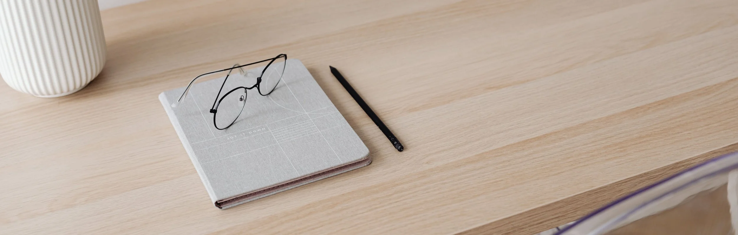 A pair of black-rimmed glasses resting on a closed planner or notebook on a wooden desk with a black pen nearby. A white ribbed mug is partially visible on the left side.