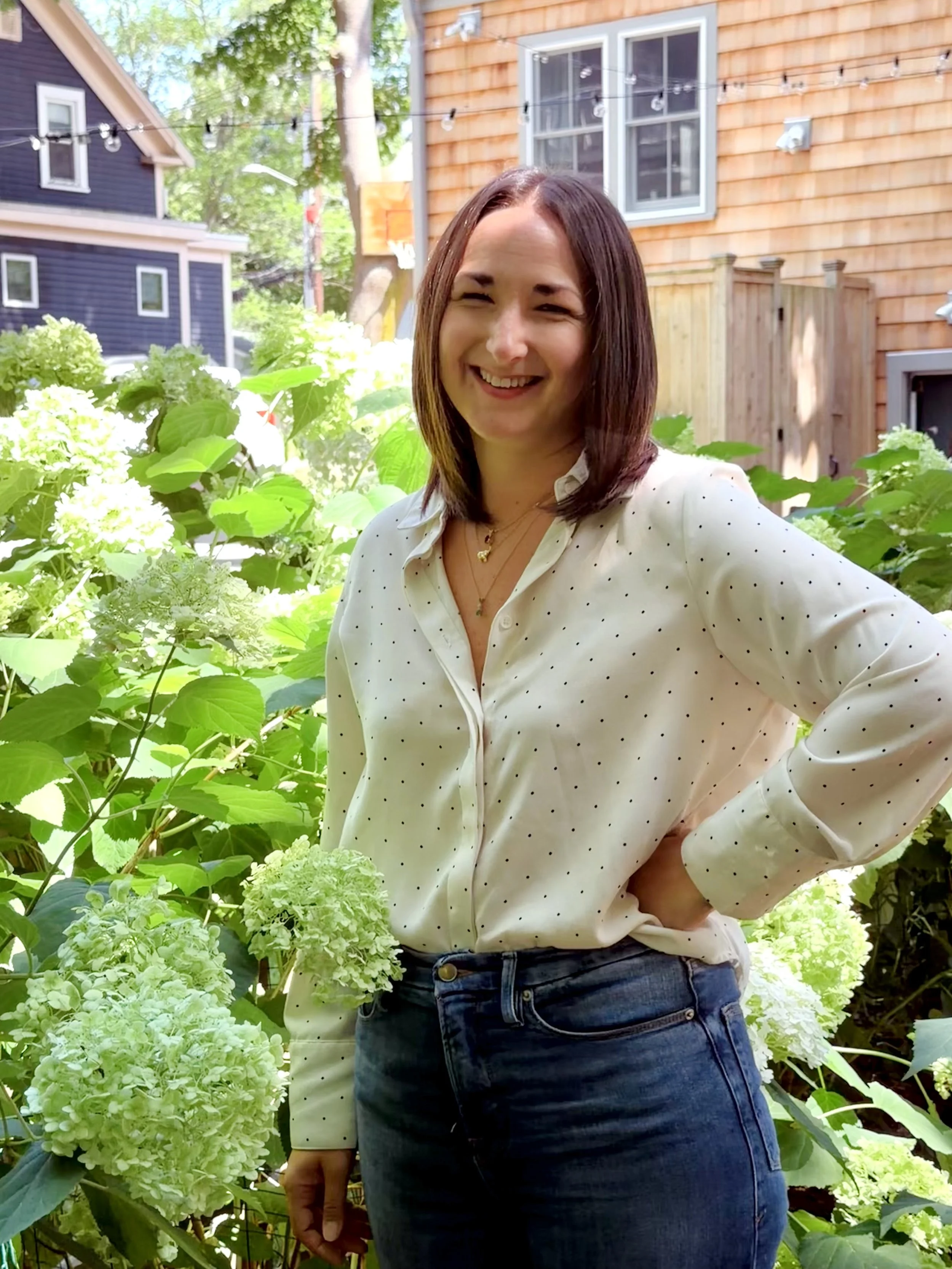 A woman with shoulder-length brown hair smiling outdoors, standing in front of green hydrangea flowers and a wooden house with a fence and windows in the background.