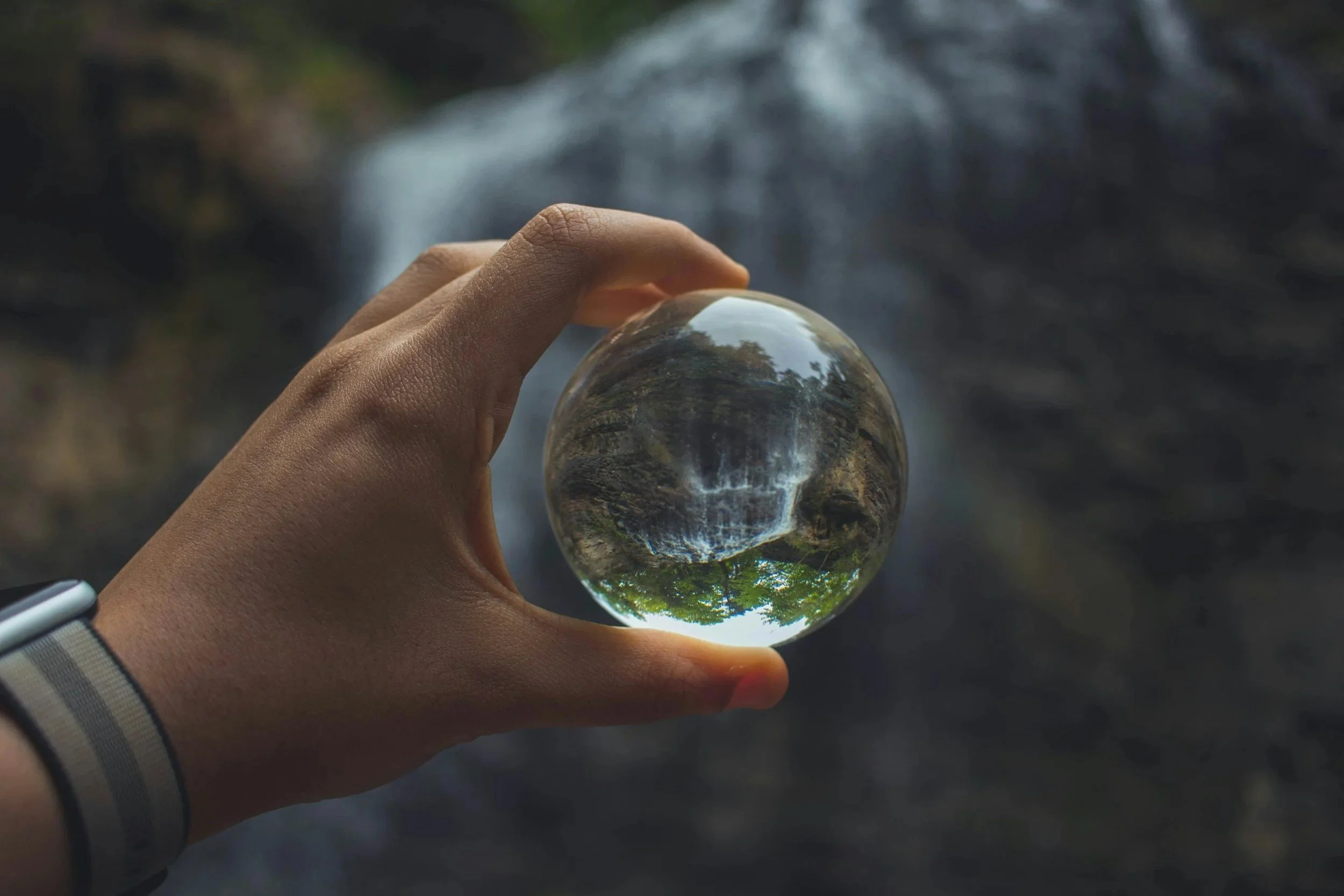 A hand holding a clear glass sphere, viewing a distorted reflection of a natural landscape with trees and water, with a waterfall in the background.