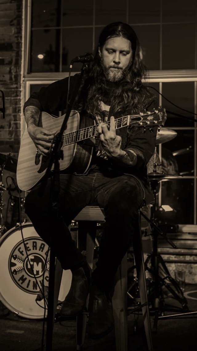 A man with long hair and a beard playing an acoustic guitar on stage at a music venue.